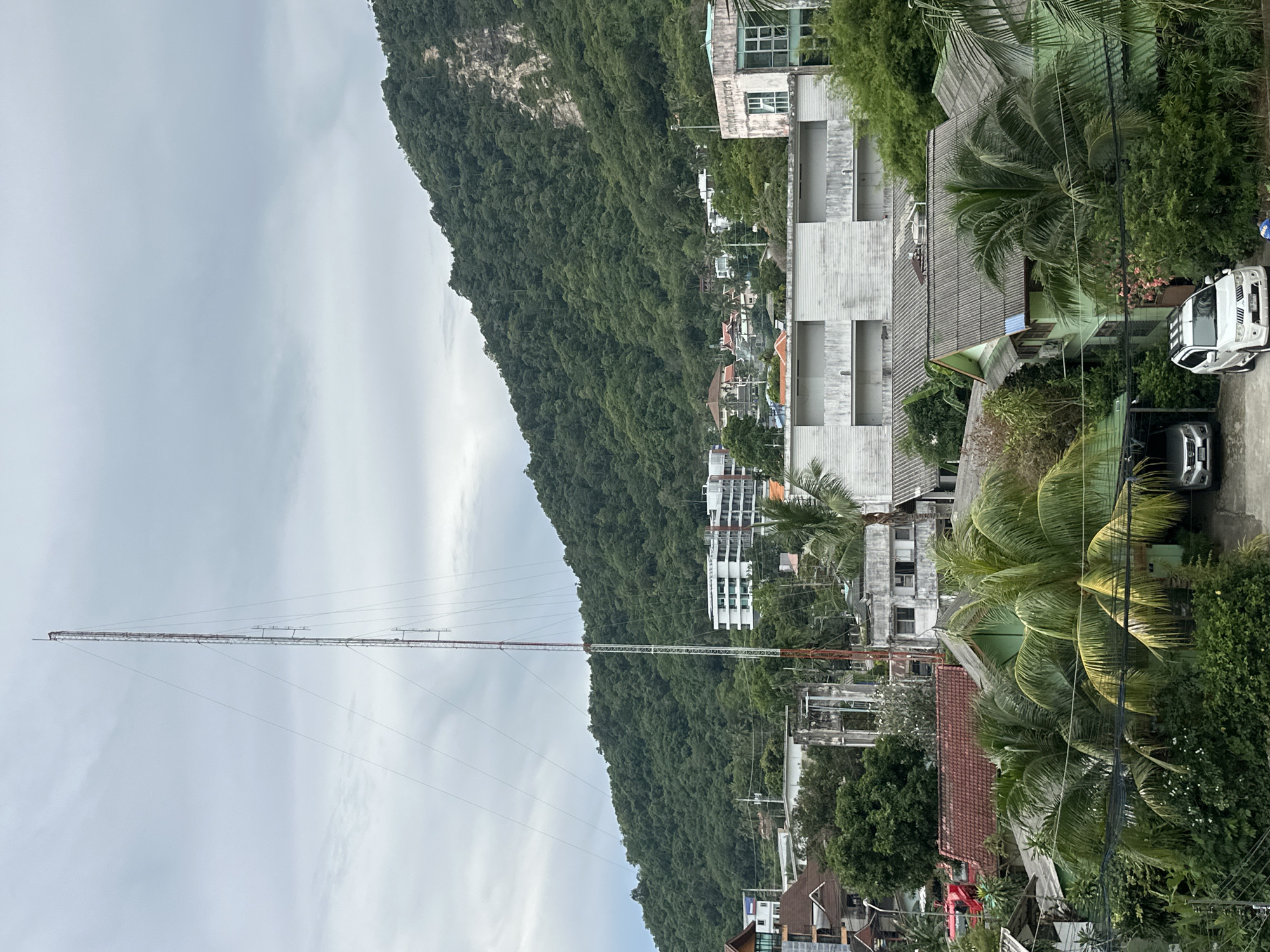 The image depicts an aerial view of Karon, Thailand, a coastal town characterized by lush greenery and a mix of residential and commercial structures. In the foreground, several houses with distinctive green roofs and palm trees are visible, hinting at a tropical climate. A white SUV is parked in front of one of the houses.
Further into the scene, a large, imposing telecommunications tower stands tall against a cloudy sky, indicating a hub for communication infrastructure within the town. Behind the tower, a densely forested mountain looms, suggesting the natural landscape surrounding Karon. The presence of modern buildings, such as a white multi-story structure with balconies, indicates ongoing development and a blend of traditional and contemporary architecture.
The overall atmosphere is overcast, suggesting a potentially humid or rainy day, common in tropical regions like Thailand. There are no visible people in the image, but the presence of vehicles and structures implies a populated area. The scene showcases the characteristic topography and urban planning of Karon, with its proximity to natural elements and evolving infrastructure.