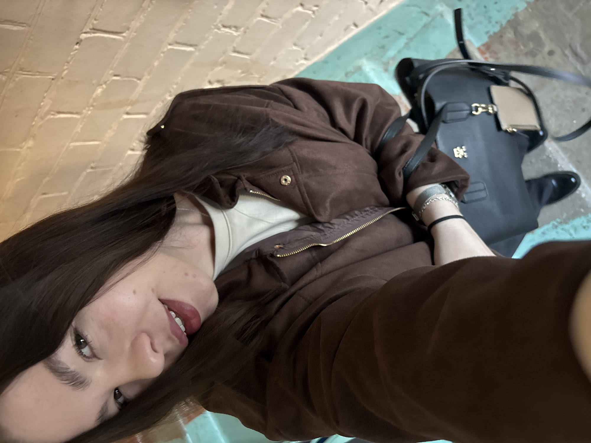 A young woman is captured in a selfie, looking directly at the camera with a slight smile. She has long, dark brown hair, brown eyes, and is wearing dark red lipstick. Her left cheek has a few small moles. She is dressed in a brown, possibly suede or velvet, jacket over a light-colored top, with a golden zipper visible on her jacket. On her left wrist, she wears a silver-toned mesh bracelet and a darker, perhaps elastic, bracelet.
To her right, a black handbag or backpack with golden hardware, including a buckle and a stylized logo, rests on a surface. A light-colored tag hangs from the bag's strap, and a black shoe or boot is partially visible next to it.
The background features a light-colored brick wall in the upper left, suggesting an urban or industrial setting. Below the brick wall and to the right, there are steps or a sloped surface with visible patches of light blue or teal paint, indicating a somewhat worn or aged environment. The overall scene suggests an outdoor or semi-outdoor location, possibly in Perm, Russia, with casual daytime lighting. No discernible text is visible on any objects or in the background.
