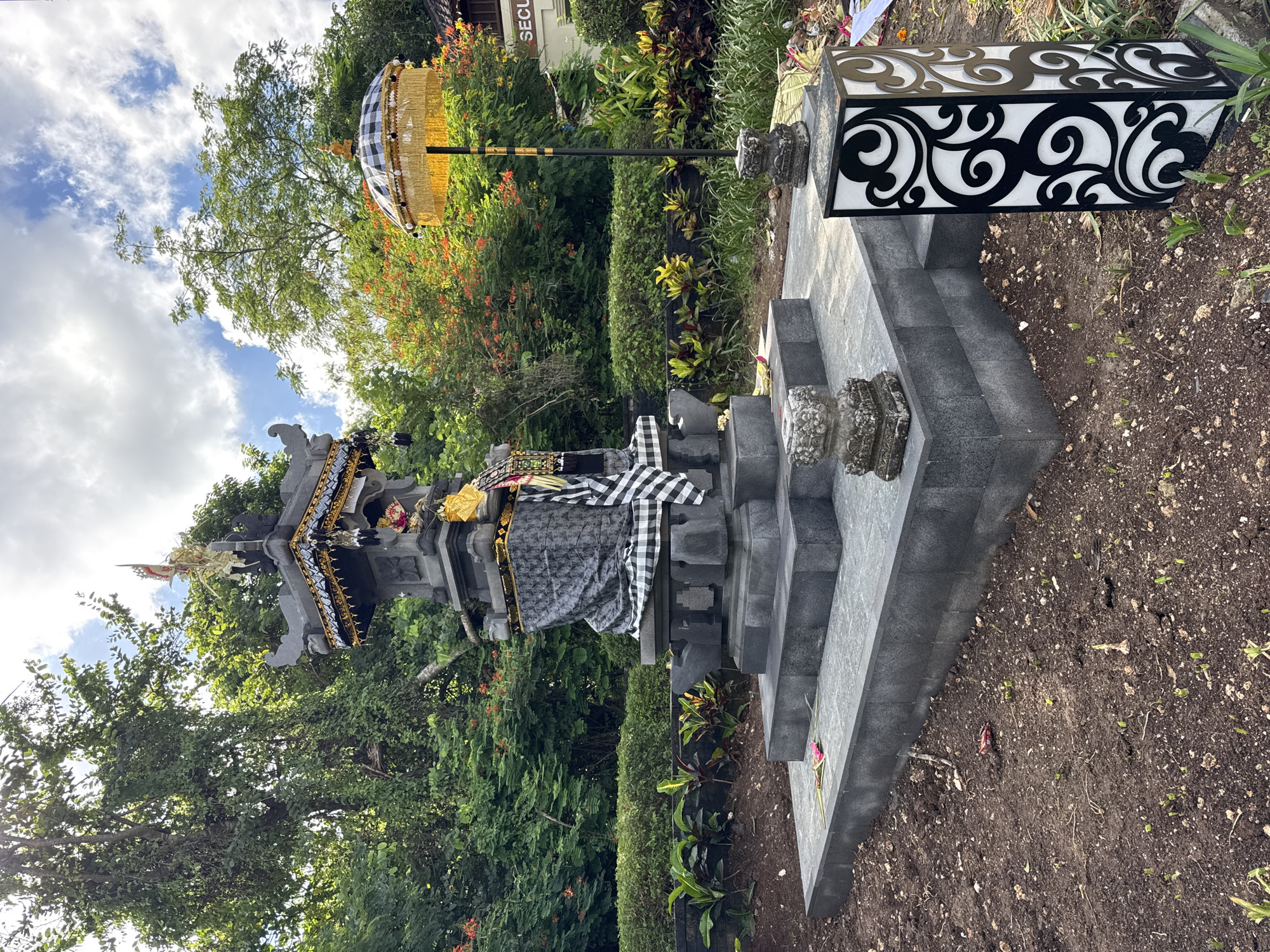 The image captures an outdoor scene in Kuta Selatan, Indonesia, featuring a traditional Balinese temple shrine and ceremonial elements under a bright, partly cloudy sky.
In the center, a multi-tiered stone shrine stands on a stepped platform of dark grey stone. The shrine is intricately carved with traditional Balinese motifs and adorned with golden and black decorative elements. A black and white checkered cloth (poleng) is draped over a portion of the shrine, a common Balinese offering or symbolic covering. Gold and white decorative items, possibly offerings or regalia, are placed on top.
To the right of the shrine, a tall ceremonial umbrella (tedung) rises, featuring a black and white checkered pattern on its canopy, fringed with golden fabric. The umbrella is supported by a dark pole. Further to the right, on a lower stone platform, rests a rectangular decorative object resembling a modern lantern or light box. It has white panels with bold, intricate black swirling patterns.
The setting is lush with greenery. Large trees with dense foliage and various bushes and smaller plants, some bearing red flowers, form a vibrant background. The ground immediately surrounding the stone structures is dark, bare earth, suggesting a garden bed. In the upper background, partially visible through the trees, is a white building with text that appears to read "SECU", likely part of "SECURITY". The overall impression is of a well-maintained, culturally significant outdoor area, basking in clear daytime sunlight.