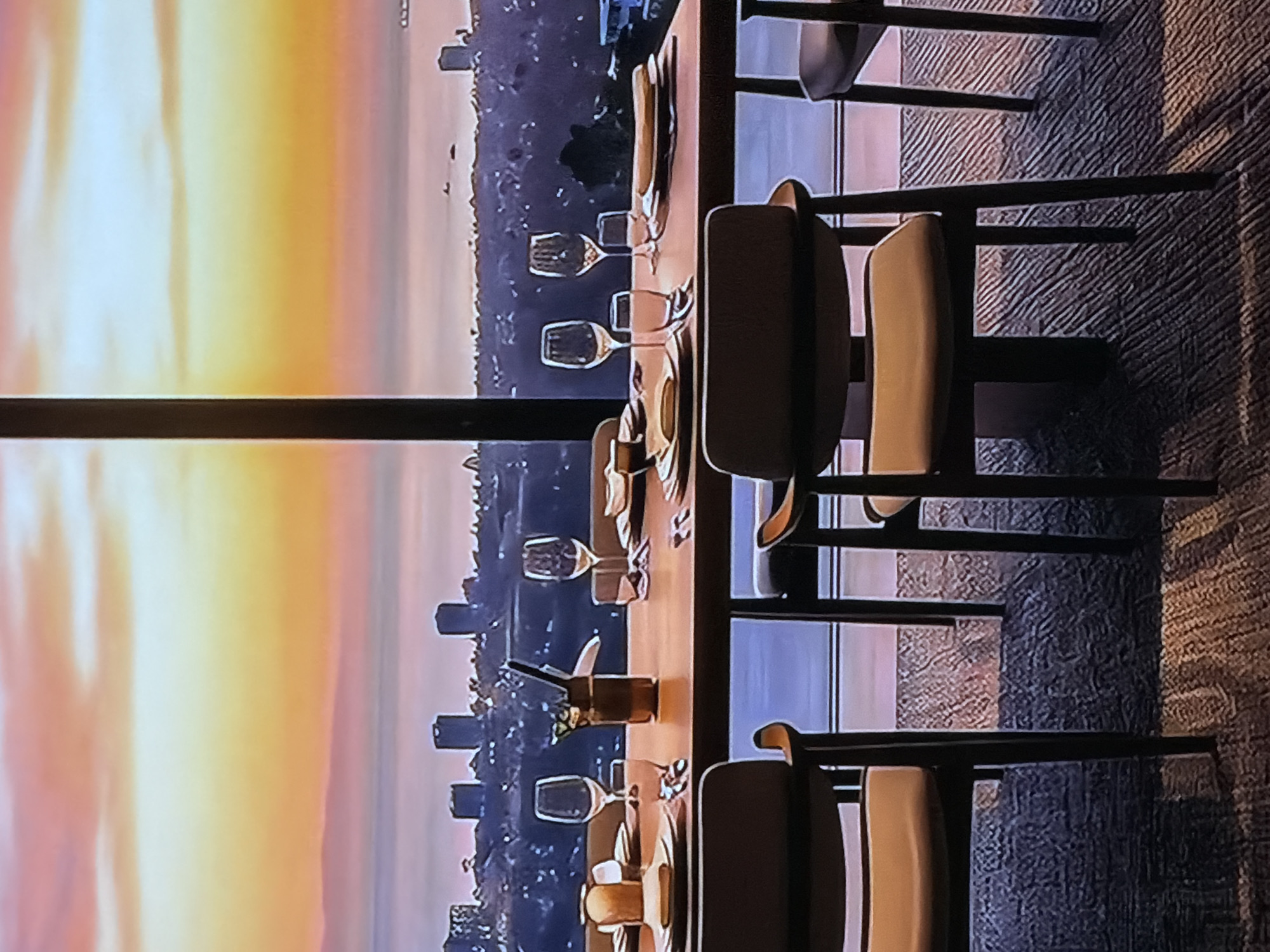 The image captures an elegant dining room interior with a panoramic view of a city skyline at sunset. The photograph appears to be taken from inside the room, looking outwards.
In the foreground, a long, polished wooden dining table is meticulously set for multiple guests. Each place setting includes plates, cutlery (forks and knives are visible), and an assortment of wine and water glasses, suggesting an upscale dining experience. Small, dark decorative items or condiments are sparsely placed along the table's center. Modern dining chairs with dark frames and light, possibly beige or cream-colored, upholstered seats and backs are arranged around the table. The floor is carpeted with a textured, patterned weave in hues of brown and grey.
A large, possibly floor-to-ceiling, window separates the interior from the exterior view. A prominent, dark vertical support beam from the window frame is visible. Beyond the window, a breathtaking sunset illuminates the sky with vibrant yellows, oranges, and pinks blending into softer purple and blue tones near the horizon. Below the horizon, a sprawling cityscape unfolds, featuring numerous buildings and structures, some of which appear to be high-rises. There are also darker areas of what might be forested land or water within the city view.
The time of day is clearly sunset, creating a dramatic contrast between the warm, bright exterior and the more subdued interior lighting. No people are visible, emphasizing the readiness and anticipation for an event or meal. The overall atmosphere is one of sophistication and tranquility, set within a high-rise building. While the provided location context is Sofiivska Borshchahivka, Ukraine, there are no specific identifying textual or landmark cues within the image itself.