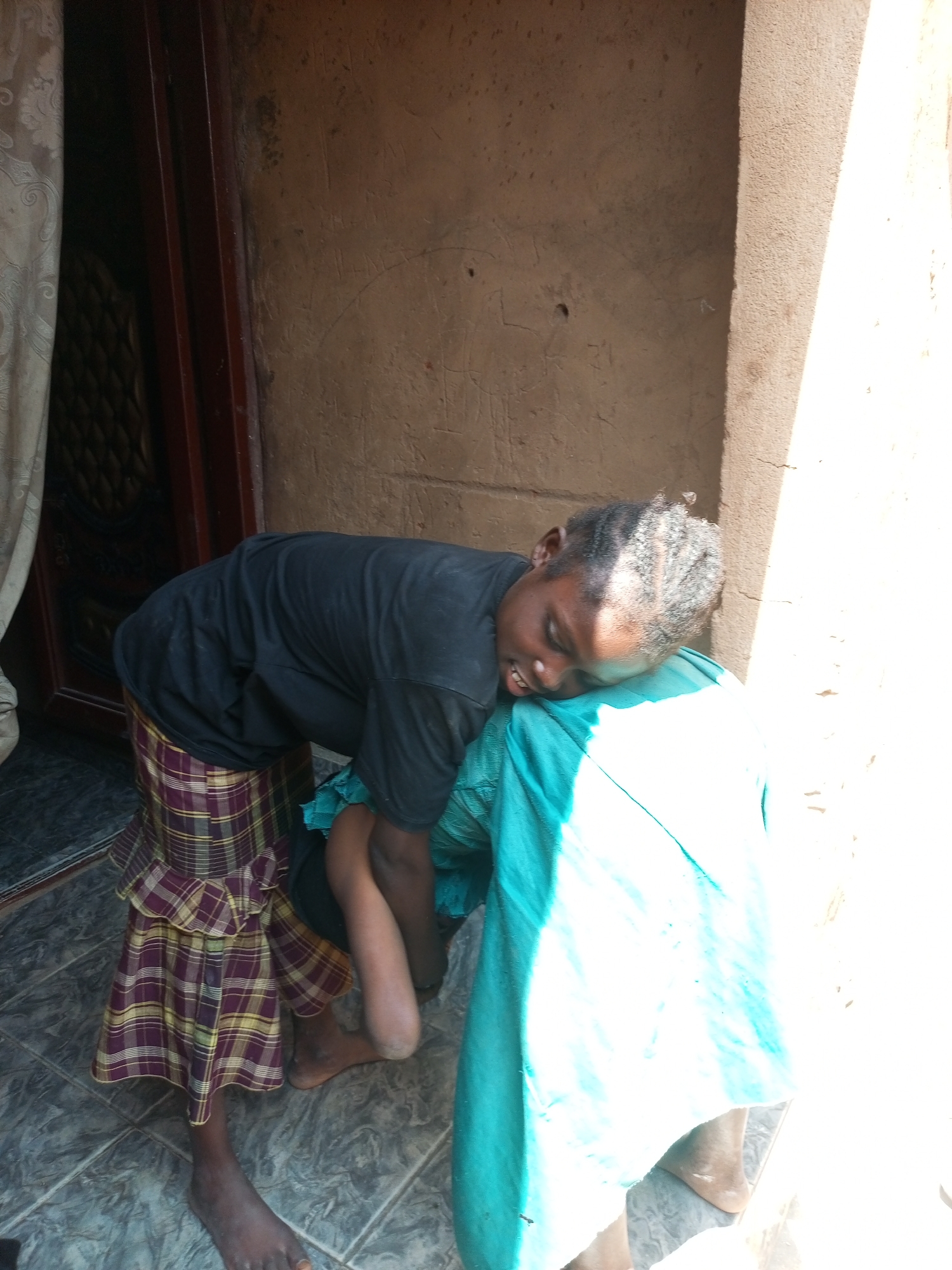 A young Black person wearing a black t-shirt and a plaid skirt is leaning over a bright blue cloth, with their head resting on it. Their eyes are closed, and their mouth is slightly open. The person's hair is styled in braids. Their bare feet are visible on a tiled floor. In the background, a rough, textured wall is visible, along with a wooden door frame and a curtain. The scene is illuminated by bright sunlight. The image appears to be taken outdoors, possibly in Tempure, Nigeria.