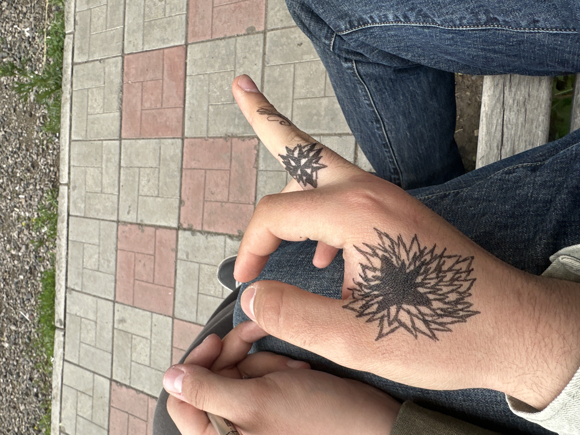The image presents a close-up, overhead view of a person's lap and hands in an outdoor setting. The individual is wearing blue denim jeans, visible across the upper thighs and knees, suggesting they are seated, possibly on a wooden bench, a part of which is visible as a grey, textured plank on the right.
The primary focus is on two hands. The upper hand, positioned on the left thigh, prominently displays two temporary tattoos drawn with black ink. A larger, intricate, star-like or floral design with numerous spiky petals covers the back of the hand. A smaller, similar design is placed on the base of the thumb. The fingers of this hand are slightly bent, and the skin shows faint hair. Below it, a second hand holds or rests underneath, with its fingers lightly gripping. This lower hand features neatly trimmed fingernails and a silver-colored ring on the ring finger.
The background consists of a patterned paved area made of interlocking rectangular pavers in alternating light grey and reddish-brown tones. To the far left, beyond the paving, there is a gravelly ground with scattered patches of green grass. The lighting suggests it is daytime. There is no visible text in the image.