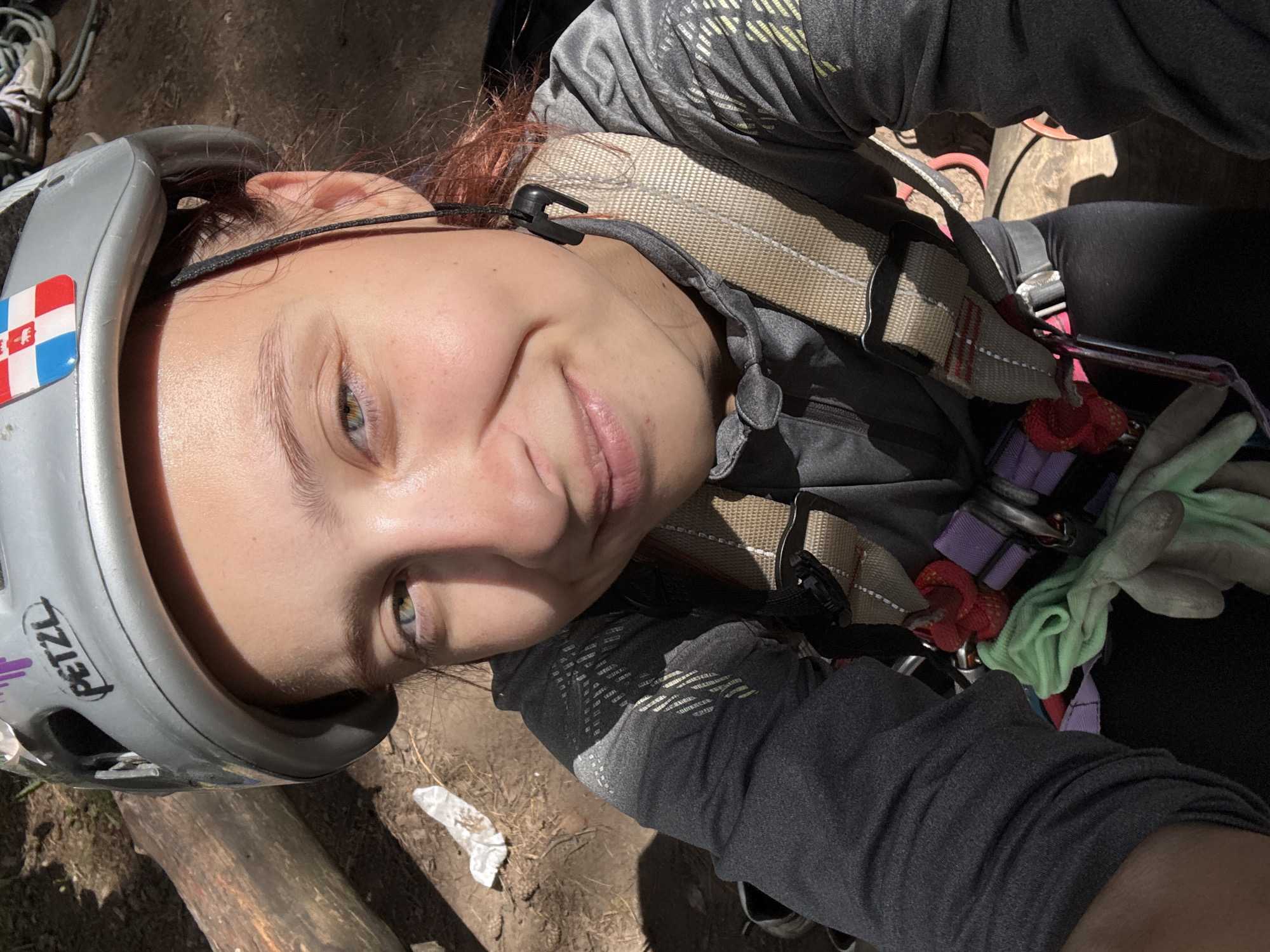 A young woman with light brown hair and green eyes smiles at the camera. She is wearing a silver climbing helmet with a PETZL logo and a flag sticker on the front. A black lanyard hangs from the helmet. She is also wearing a grey long-sleeved shirt and a climbing harness with tan straps and various colored gear attached, including a red rope and a green glove. The background appears to be outdoors with a log and dirt visible. The lighting suggests it is daytime. This scene is not identifiable as being in the city of Chaikovsky, Russia.