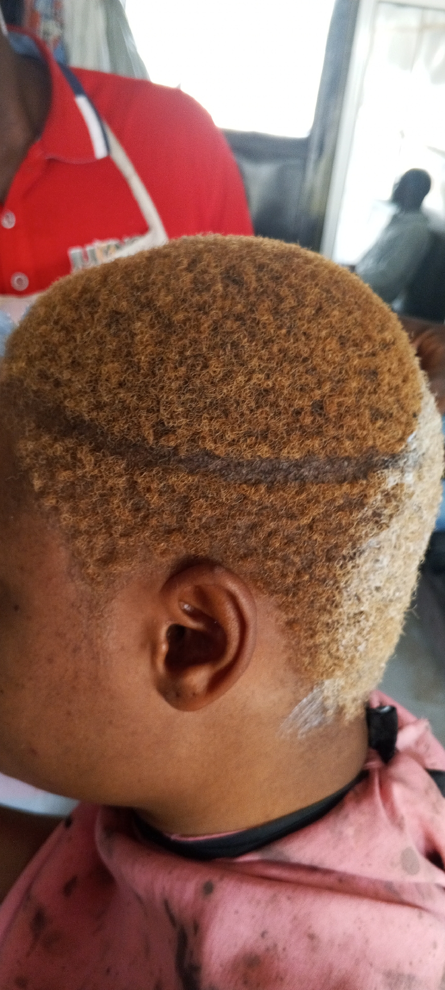 The image shows a close-up view of the side of a person's head, focusing on their short, textured, light brown hair. A dark line is visible across the top of the head, potentially marking a section for a haircut or style. White product, possibly hair dye or styling product, is applied to the lower right side of the hair and down the neck. The person's ear is also visible, and their skin appears to be dark.
In the background, a person wearing a red polo shirt with white and blue trim is partially visible on the left. To the right, a blurred background suggests an indoor setting, possibly a vehicle or a room with a window. The lighting appears to be bright, suggesting it could be daytime. There is no visible text in the image. The context provided is Jalingo, Nigeria.