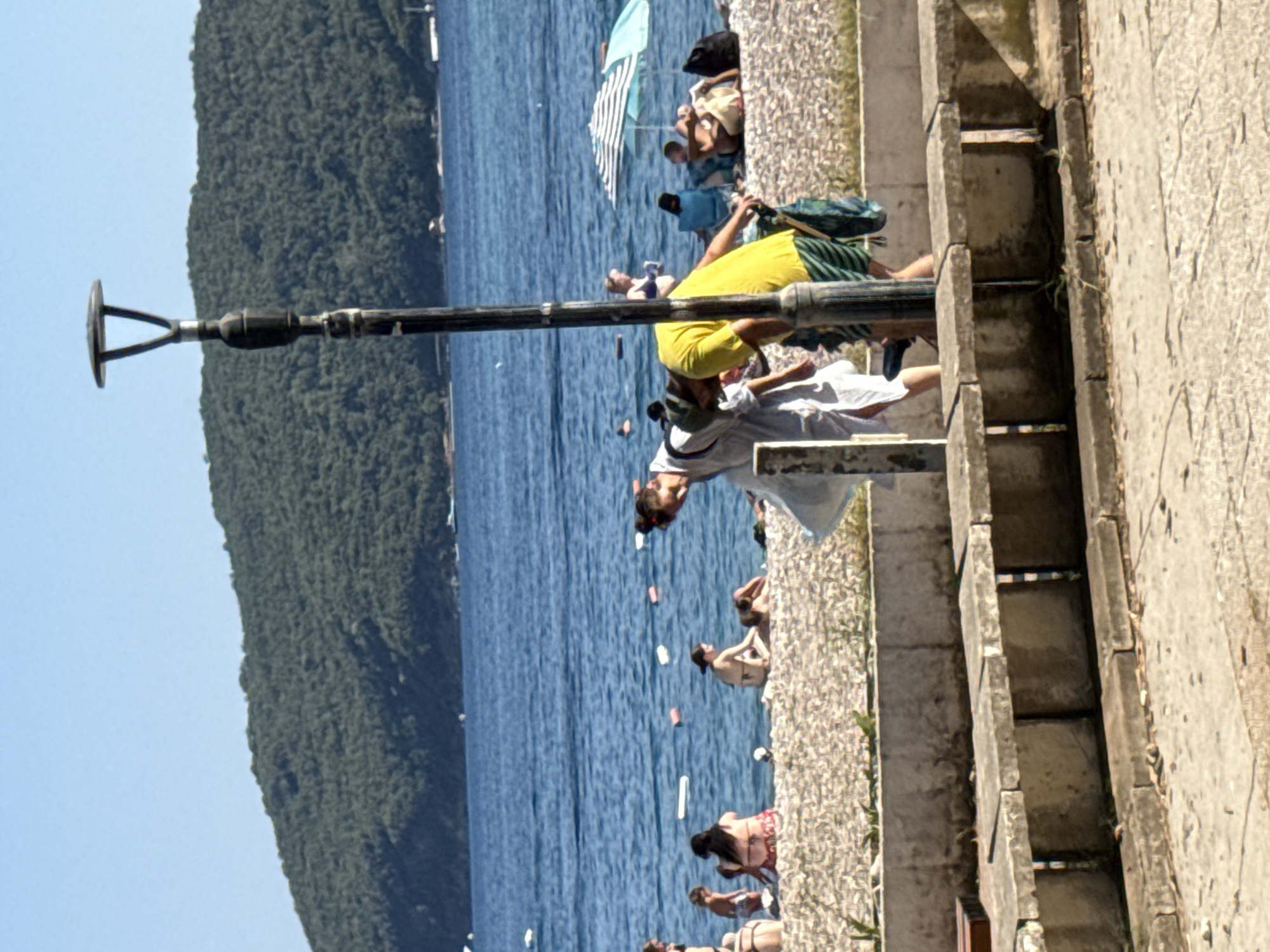 The image shows a view of the beach in Budva, Montenegro, with the sea, a forested hill, and people. In the foreground, a man in a yellow t-shirt and striped shorts bends down, holding a blue bag. Next to him, a woman in a white dress also bends over. A lamppost stands between them, obscuring part of their bodies. In the background, several people are seen on the pebble beach, some sitting and others walking. The sea is calm and blue, with buoys and small boats visible in the distance. A large, forested hill forms the backdrop, characteristic of the Budva coastline. The overall scene depicts a typical summer day at the beach.