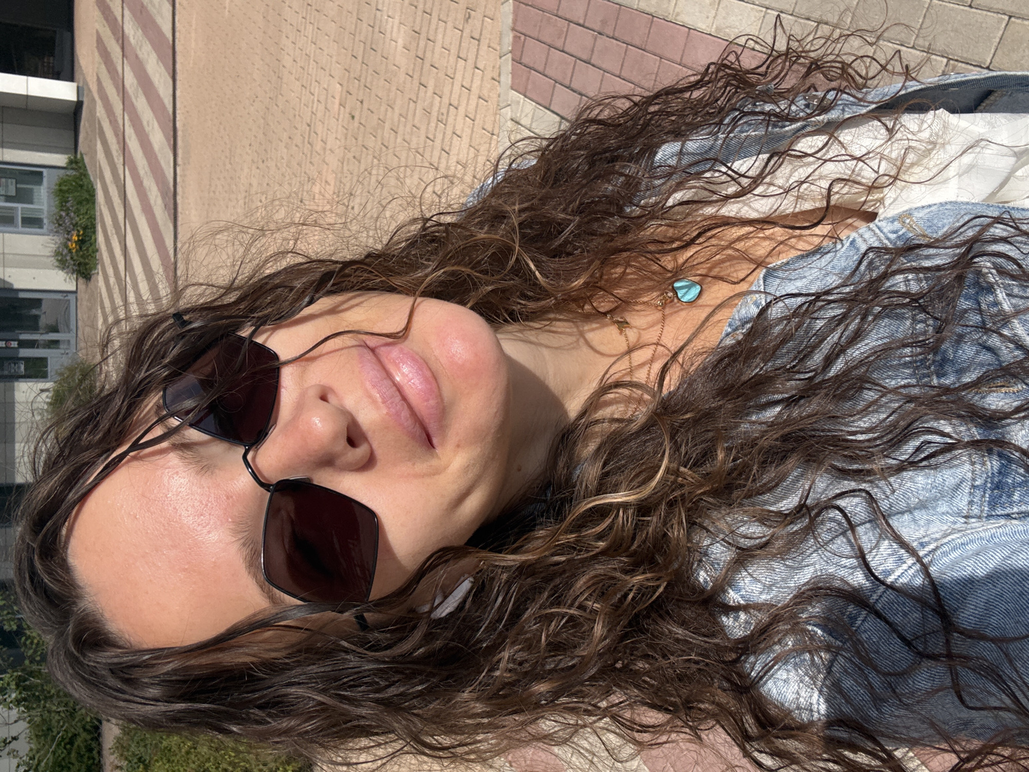 A medium shot shows a young woman with long, curly brown hair and sunglasses taking a selfie outdoors. She is wearing a white top and a denim jacket, with a turquoise heart-shaped pendant necklace. The background is an urban environment with a sidewalk made of brick pavers and a building in the distance. The woman appears to be in a public area, possibly a park or street, under natural lighting.
