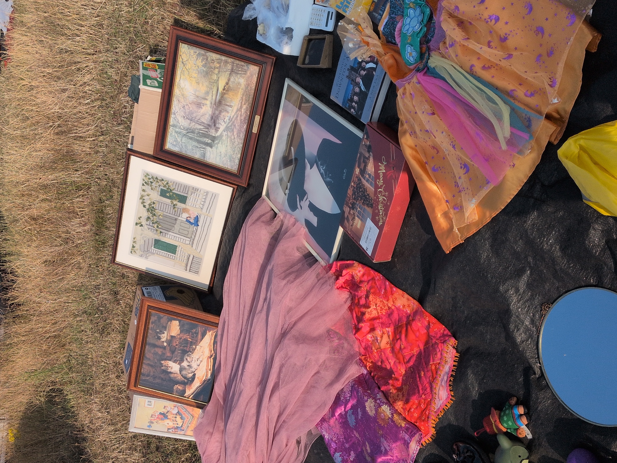 The image depicts a flea market or yard sale at a grassy outdoor location. Several framed pictures are laid out on a black tarp, including one with cats, another of a house with a green door, and a landscape painting. Various textiles, including a pink tulle skirt and brightly patterned fabrics, are also visible. A red box, a book titled "Chronicles of Downton Abbey," and a calculator are among the other items. A child's orange dress with rainbow-colored tulle accents is displayed, and a small toy figurine is also present. A blue mirror and a yellow plastic bag are also on the tarp. The background consists of dry grass and a hint of trees, suggesting an outdoor setting in a rural or semi-rural environment. The location is identified as Claverley, United Kingdom.