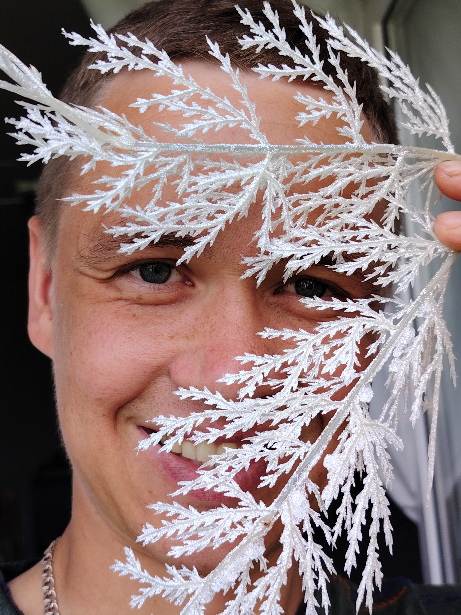A middle-aged man with fair skin, short brown hair, and blue eyes is smiling at the camera. He is holding a decorative, artificial white branch with a textured, frost-like finish in front of his face, partially obscuring his features. The man has some freckles visible on his cheek. A silver chain is visible around his neck. The background is dark on the left side, suggesting an interior space, while the right side shows a window with some light coming through. There is no visual evidence to confirm the location as Chaikovsky, Russia.