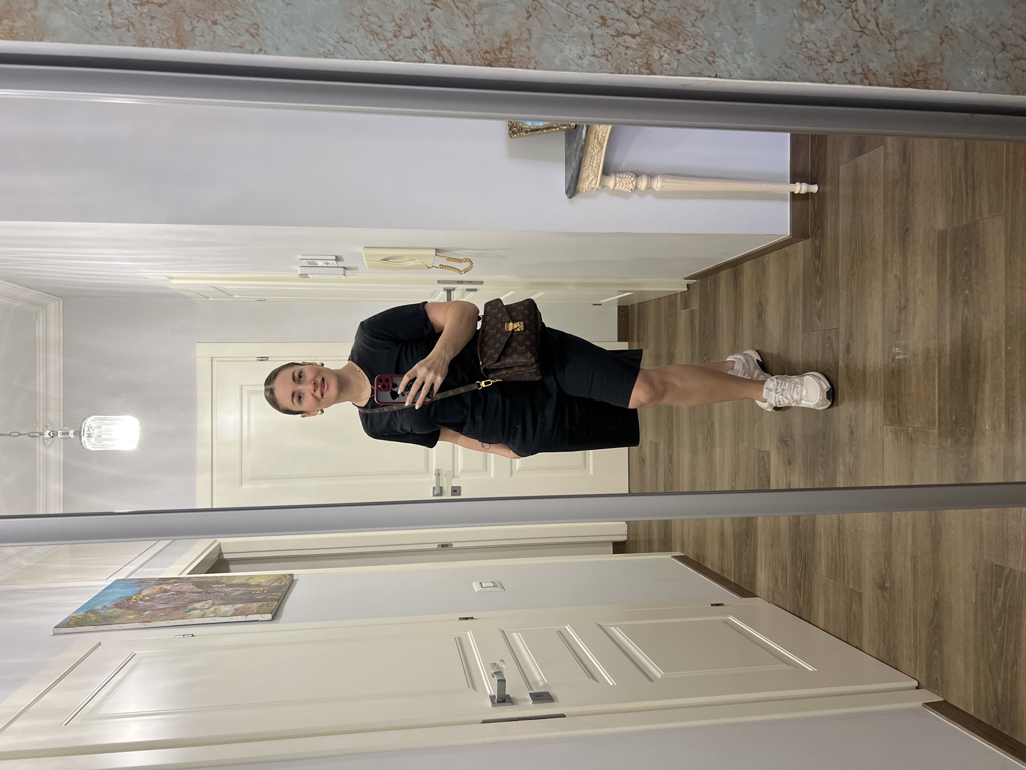 A woman in her late 20s or early 30s is taking a mirror selfie in a hallway, with a white door and wood-paneled walls visible. She is wearing a black t-shirt, black shorts, and white sneakers. She has a brown designer handbag slung across her body and is holding a smartphone. The hallway features a framed painting on the wall and a decorative console table with a framed picture in the background. The flooring is made of dark wood. The image appears to be taken indoors, likely in a residential setting. The specific location in the city of Rome, Italy cannot be determined from the provided image.