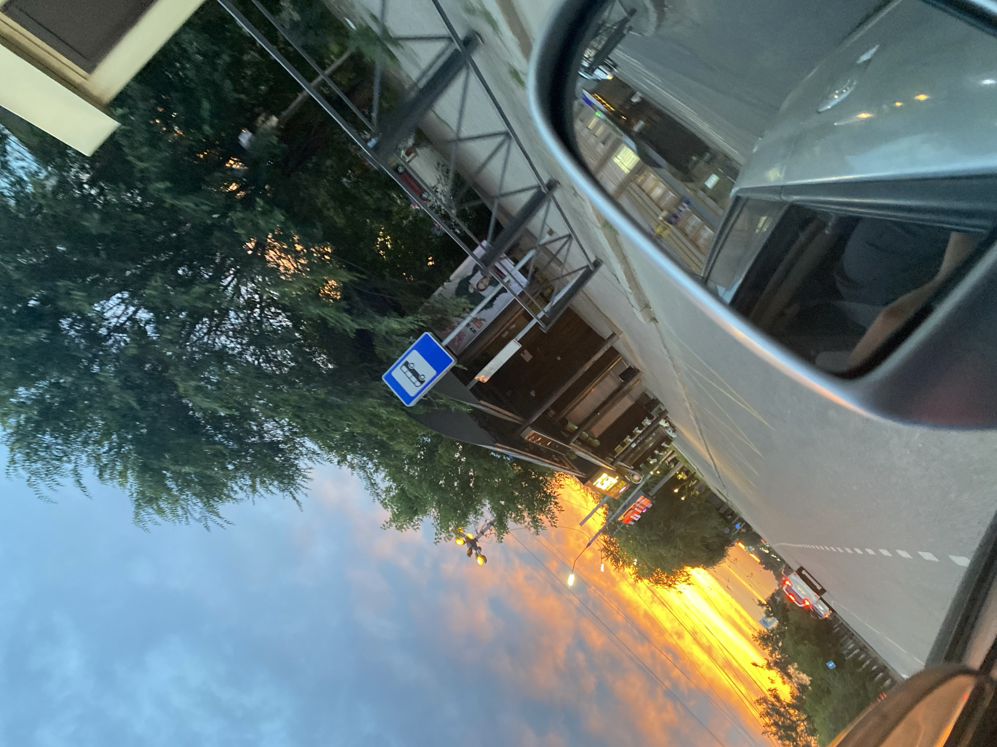 The image depicts a street scene in Irkutsk, Russia, during a vibrant sunset. The sky is filled with dramatic orange and yellow clouds, indicating the sun is low on the horizon. A road runs through the scene, with a few cars visible, including one with flashing red lights, suggesting it might be an emergency vehicle.
On the right side of the road, there is a bus stop with a blue sign indicating bus service. Adjacent to the bus stop is a building with multiple storefronts. Trees line the street, their foliage casting shadows.
The perspective of the image is from inside a vehicle, with the side mirror and a portion of the car's interior visible in the foreground. A person's arm and shoulder can be seen through the open car window. The overall impression is that of a journey through the city as daylight fades.