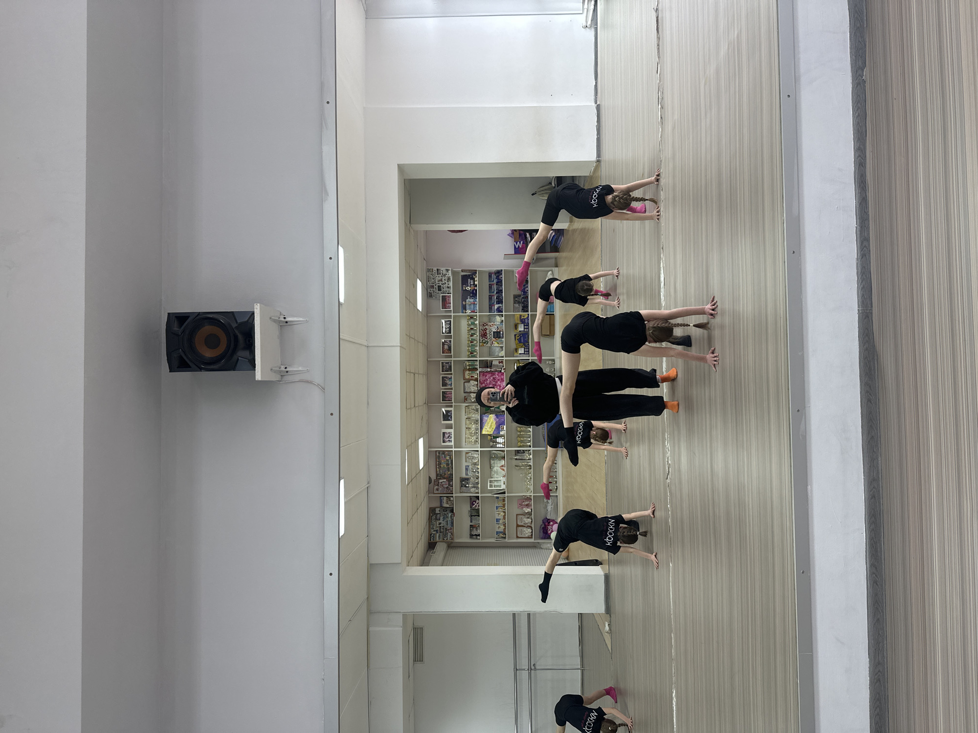 A reflective surface captures a scene of individuals in a dance or exercise studio. Six people are present, likely a mix of adults and children, all dressed in dark attire. They are engaged in a stretching or warm-up exercise, assuming a posture where their hands are on the floor and bodies angled upwards.
The background reveals a studio setting with a display case filled with trophies, pictures, and various items. The walls are white, and there is a speaker mounted on one wall. The floor appears to be light-colored.
Given the details, the location is likely a dance or fitness studio within the city of Votkinsk, Russia. The people are participating in a class or training session.