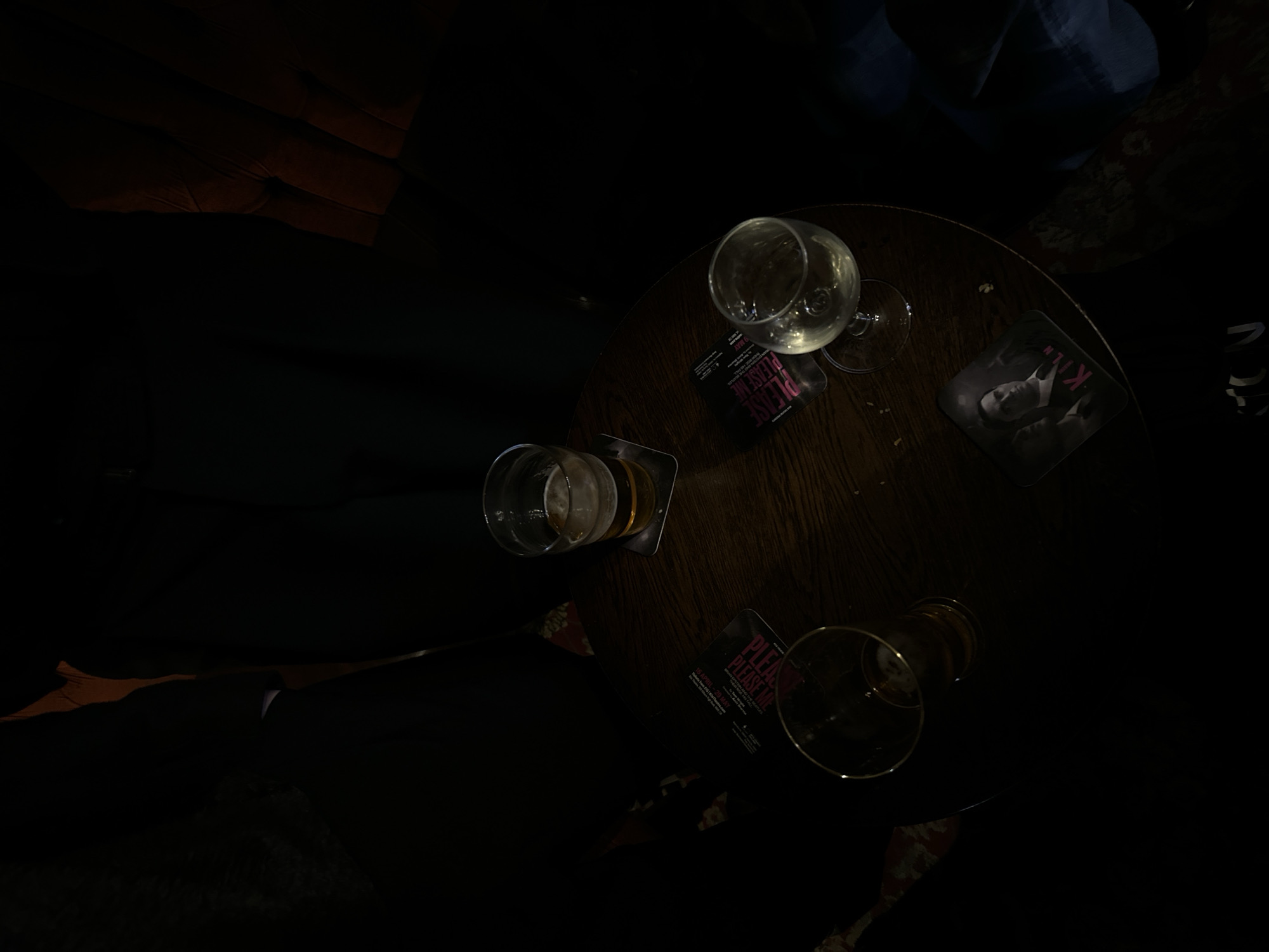The image presents a dimly lit, top-down view of a round, dark wooden table in what appears to be a pub or bar, likely in London. The low lighting indicates an evening or nighttime setting.
On the table are three drinking glasses and three coasters. A wine glass, partially filled with a pale liquid (possibly white wine), is centrally placed, resting on a coaster. To its left, a beer glass or tumbler, mostly empty, also sits on a coaster. A third empty glass, likely another beer glass, is visible in the lower right portion of the table.
Three coasters provide notable details. One prominent coaster, located to the right of the wine glass, features a black and white image of a man in a suit, with the letter "K" visible. Another coaster, partially obscured under the wine glass, displays the text "PLEASE ME" in pink. The text "TON" is also visible on one of the coasters. Further text on the coasters includes dates such as "16 APRIL - 29 MAY" and "9 MAY," suggesting promotions for events or performances.
The background is predominantly dark, with blurry shapes hinting at seating or other patrons, reinforcing the social, indoor venue. The scene captures the aftermath or a pause during a social gathering where drinks have been consumed.