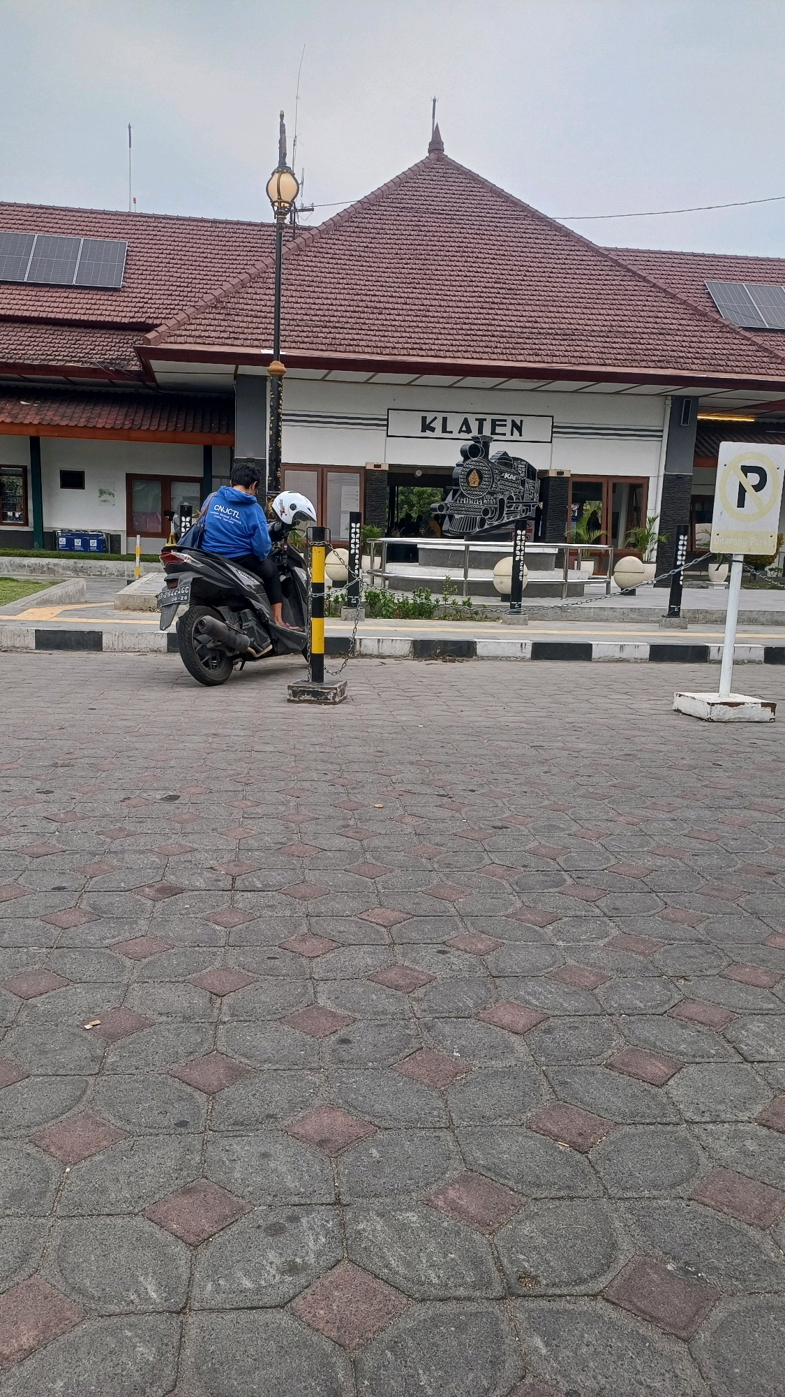 The image shows the exterior of Klaten Train Station in Klaten, Indonesia. A person wearing a blue jacket is sitting on a black scooter parked on a brick-patterned walkway. The train station building has a red tiled roof, solar panels, and a sign that reads "KLATEN". In front of the building, there is a metal sculpture of a steam locomotive. A "P" parking sign is visible on the right side of the image. The sky is overcast.