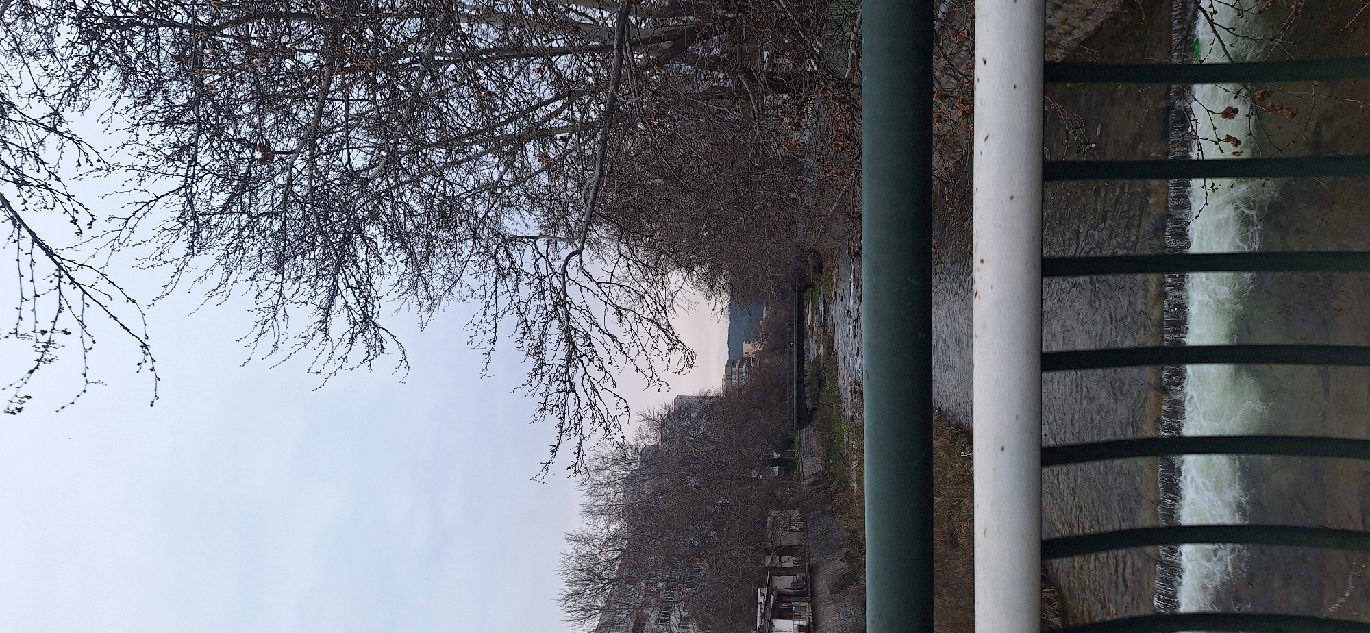 The image shows a view from a bridge in Blagoevgrad, Bulgaria, overlooking the Blagoevgradska Bistritsa River. A green and white metal railing runs along the right side of the frame. Beyond the railing, the river flows over a series of small, artificial waterfalls. Bare trees line the riverbank on both sides. In the distance, buildings are visible, with a mountain range in the background under a cloudy sky. The river is the central element, flowing from the distance towards the viewer.
