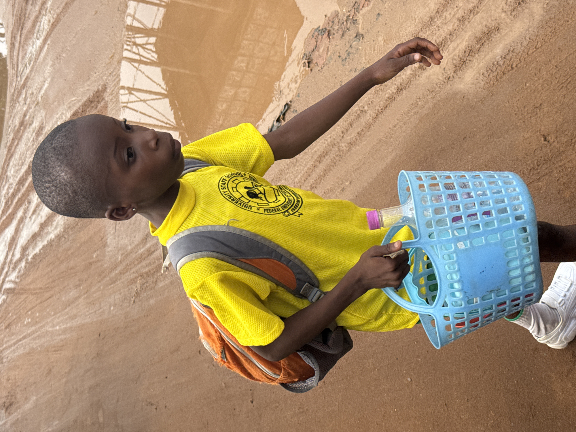 A young child, likely a boy of elementary school age, stands outdoors on a dirt or sandy path in Ihiagwa, Nigeria. The child has short, dark hair and dark skin, and is looking slightly upwards and to the right with a somewhat contemplative expression.
The child wears a vibrant yellow short-sleeved t-shirt with a grey collar. On the front of the shirt, a circular black and white logo is visible, which includes text such as "STAFF SCHOOL," "UNIVERSITY," and "FEDERAL UNIVERSITY OF TECHNOLOGY," along with the word "FREEDOM." The child is also wearing a grey and orange backpack and white sneakers with white socks.
In their right hand, the child holds a light blue, perforated plastic basket. A clear plastic bottle with a pink cap is placed inside the basket. The ground beneath the child is a light brown, earthy texture. To the left, there's a large, shallow puddle of water that reflects indistinct shapes, possibly structures or trees, from above. The overall scene suggests a sunny day, with the ground appearing dry in some areas and wet from the puddle in others, possibly indicating recent rainfall. The photograph is taken at an oblique angle, tilting the horizon.