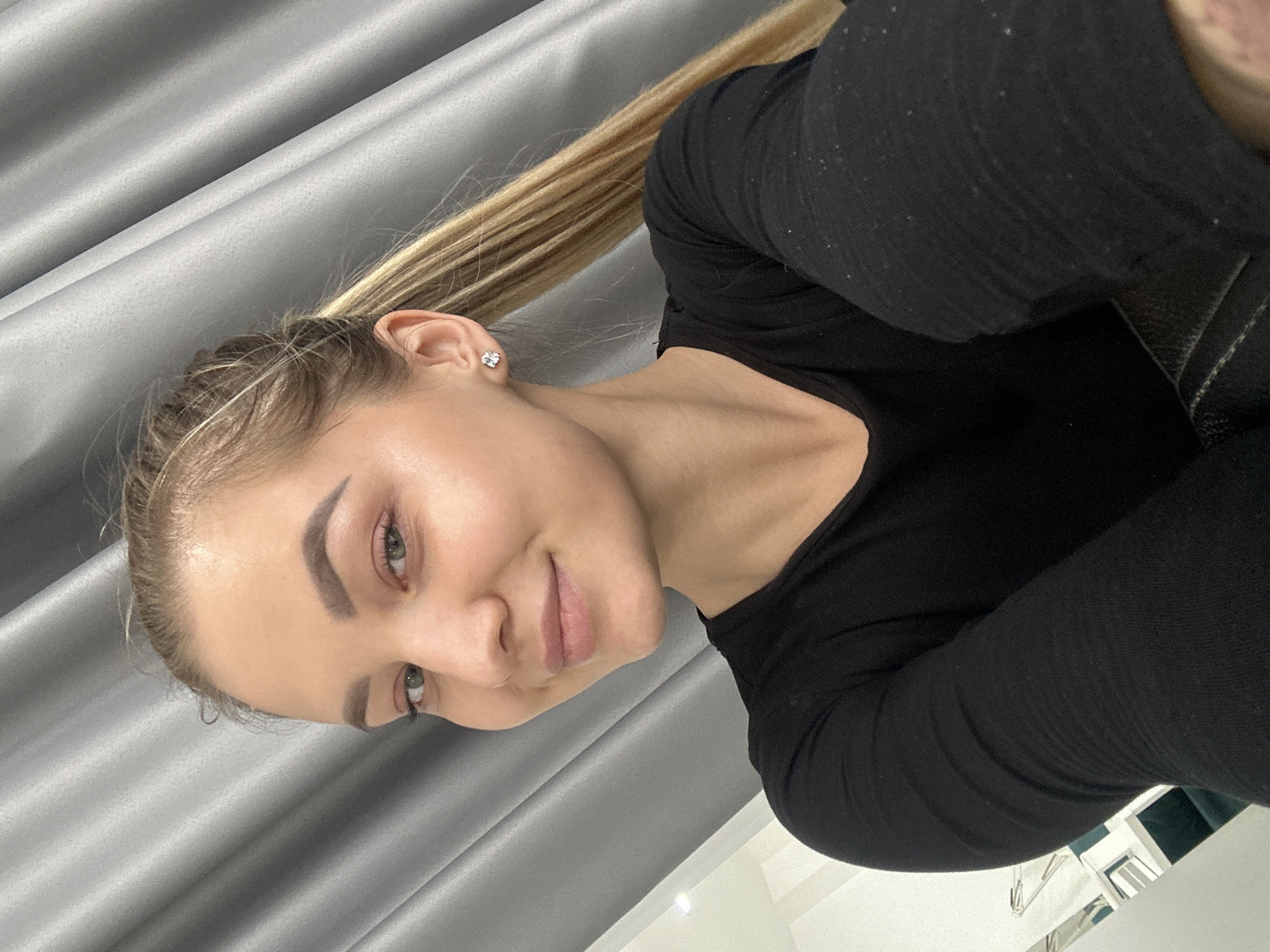 A young woman is photographed indoors. She is light-skinned, with light blonde hair pulled back into a ponytail, and is wearing a black long-sleeved shirt. She is smiling gently. A gray curtain is visible in the background. A partial view of a white object and a metallic structure is present in the lower right corner. The photograph appears to be taken from a slightly elevated, off-center angle, and is rotated counterclockwise. It is not possible to confirm that the location is Budva, Montenegro from the information available.