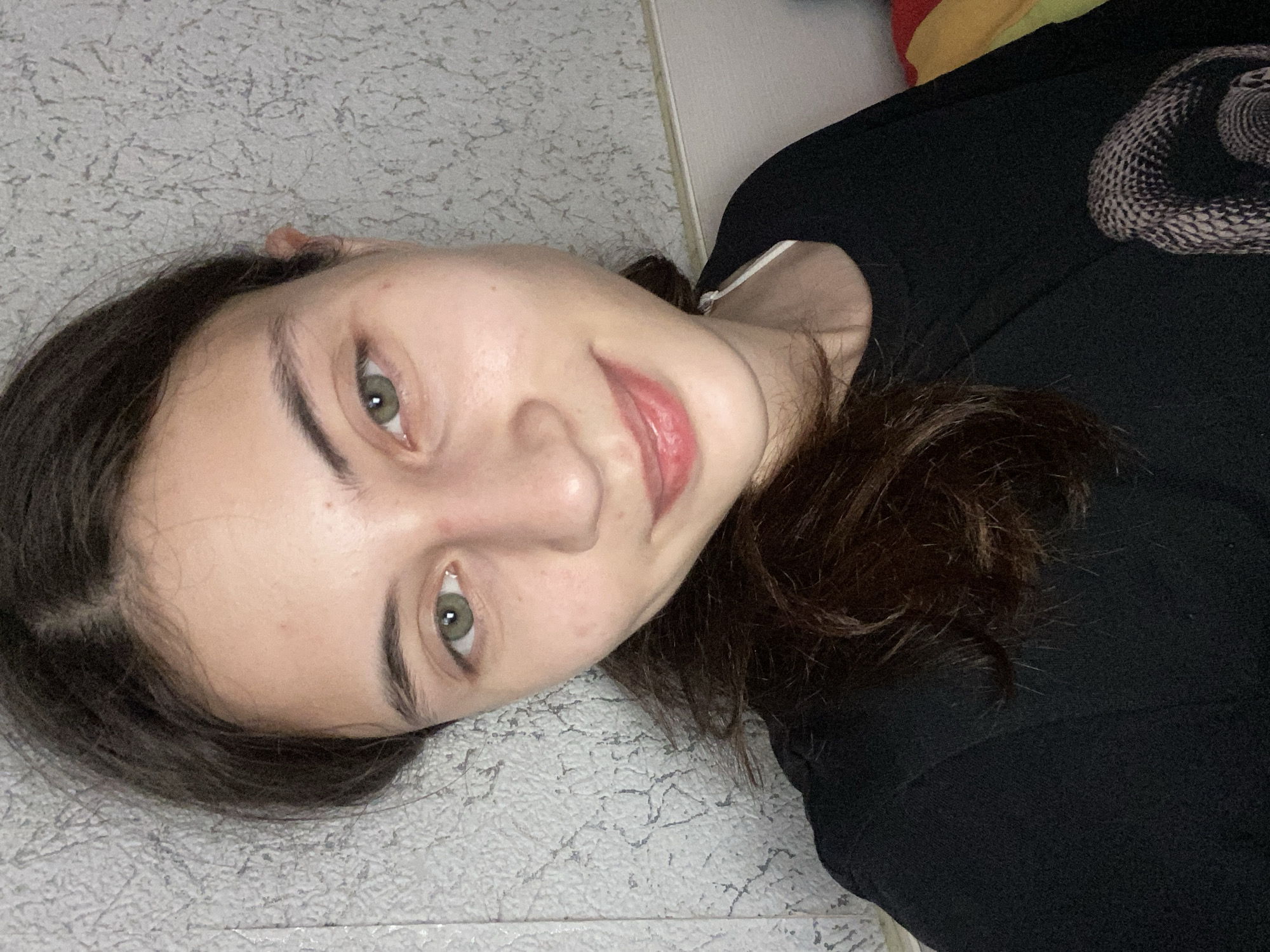 This media file is a portrait of a young person lying down, possibly indoors on a textured floor. The person has dark hair and green eyes, and is looking towards the camera with a slight smile. They are wearing a black shirt with a snake graphic on it. The lighting suggests it could be daytime, but the specific time of day is not discernible. The background is a textured gray surface, with hints of other objects or fabrics in the upper right corner, one appearing to be colorful. The overall mood is relaxed and casual. There is no visible text in the image.