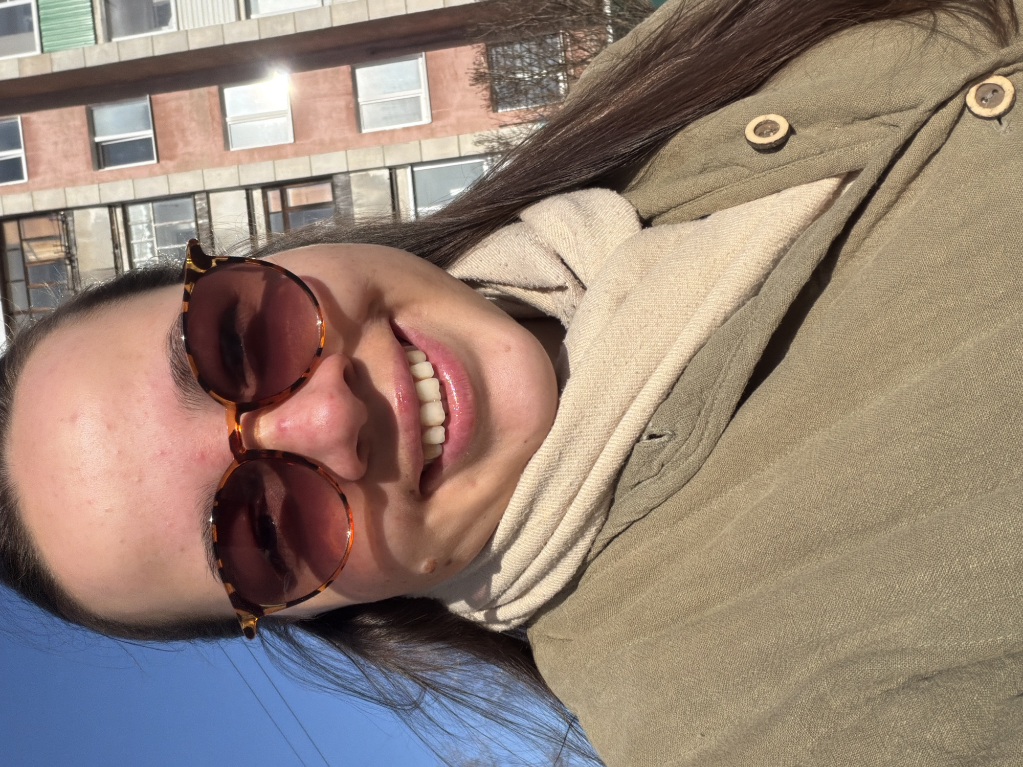 A young woman smiles and wears round sunglasses. She is wearing a light-colored scarf and a greenish-brown button-down shirt. She is outdoors on a sunny day. In the background is a building with multiple windows. The sky is blue. The location is likely in Chaikovsky, Russia.