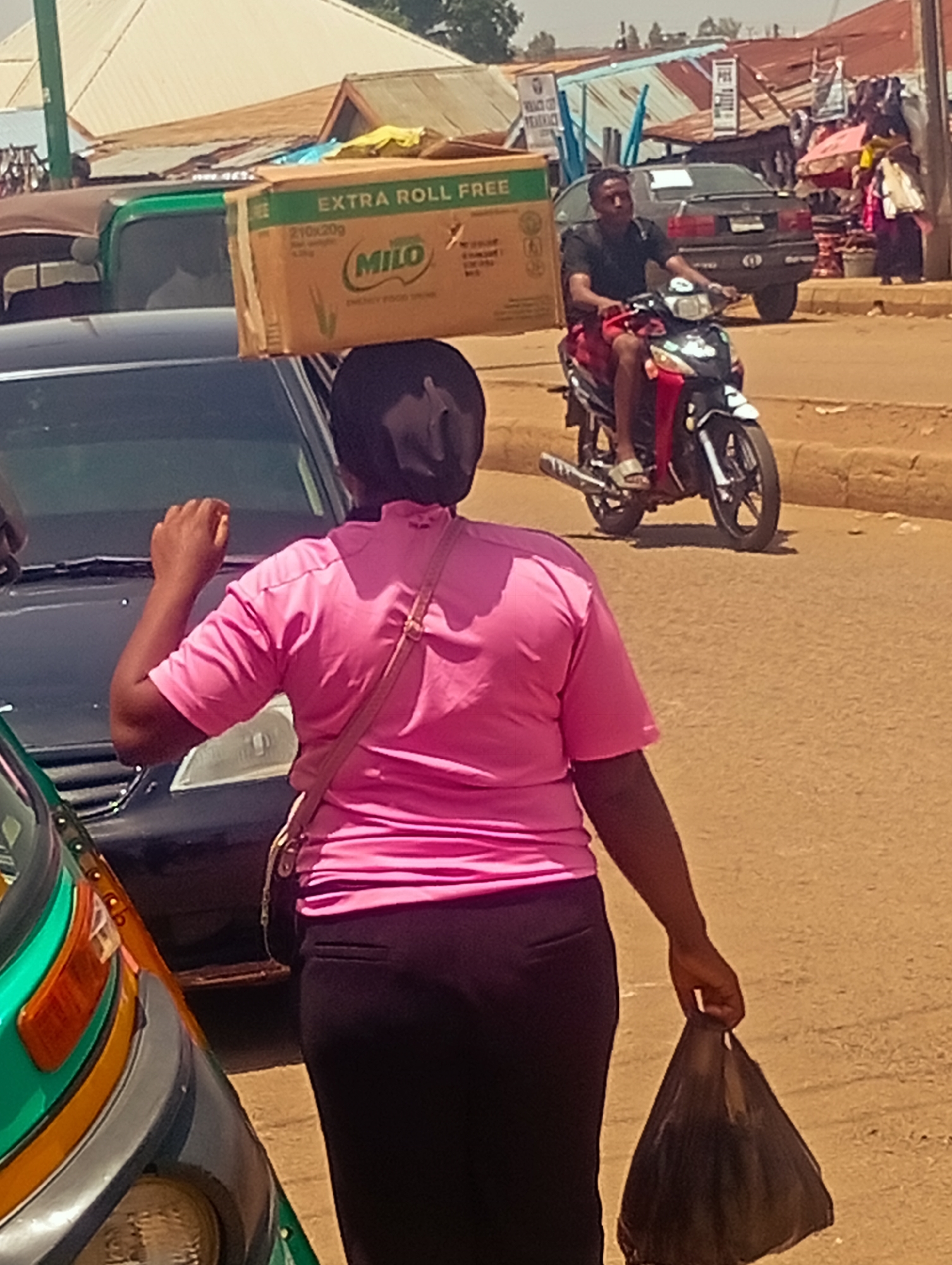 In Bukuru, Nigeria, a person is seen from behind walking with a large cardboard box labeled "EXTRA ROLL FREE" and "MILO ENERGY FOOD DRINK" balanced on their head. The person is wearing a bright pink t-shirt and dark pants, with a light pink strap bag crossing their chest. They are carrying a black plastic bag in their right hand. The scene takes place on a dirt road with other vehicles present, including a black car and the front of a green and orange auto-rickshaw. In the background, a man is riding a motorcycle, wearing a black t-shirt and red shorts. Buildings with corrugated metal roofs and various stalls are visible, suggesting a marketplace or commercial area. The environment appears dry and sunny.