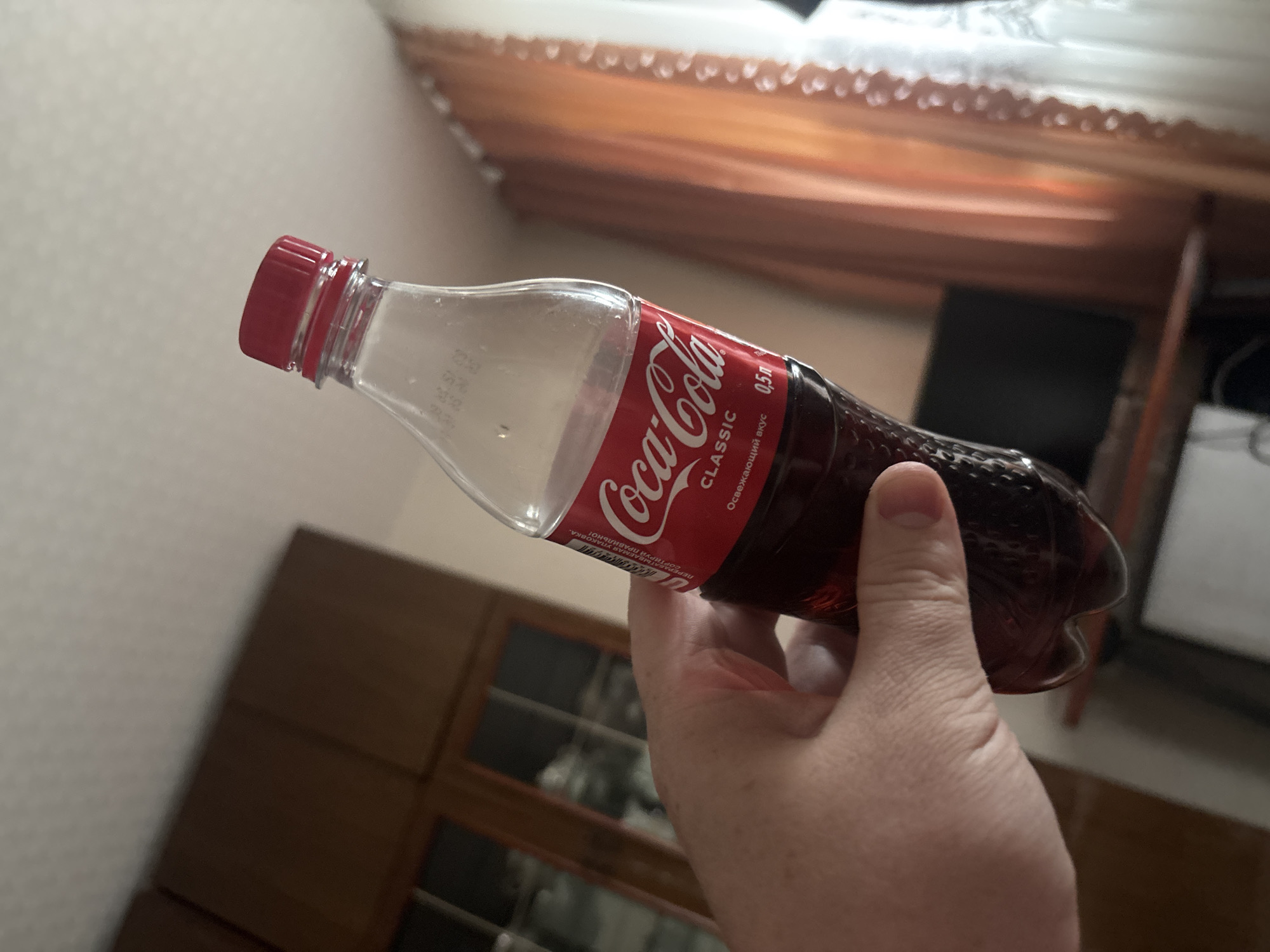 A hand is holding a clear plastic bottle of Coca-Cola Classic. The bottle is half-full of dark brown liquid and has a red cap. The label on the bottle is red and white and features the Coca-Cola logo. The background is blurry and appears to be an indoor setting with wooden furniture and curtains. There are no people visible other than the hand holding the bottle.