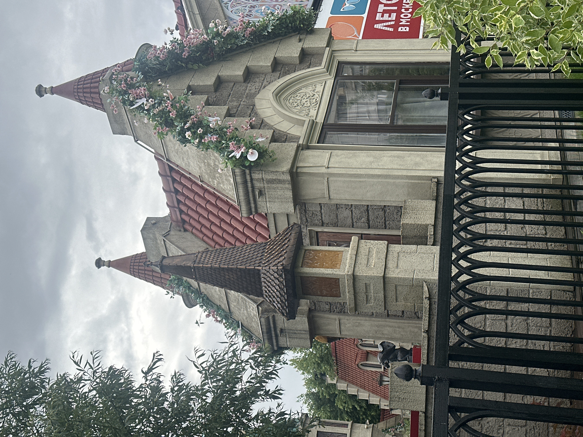 The image shows the facade of a whimsical, castle-like building, possibly a commercial establishment or a themed attraction, in Moscow, Russia. The architecture features pointed turrets with red tile roofs, stone-like cladding, and decorative floral garlands with paper birds. A black wrought-iron fence with Gothic-inspired arches runs across the foreground, partially obscuring a stone wall and a small statue of a gargoyle-like creature. To the right, a red and white advertisement banner with text "ЛЕТО" (Summer) and "В МОСКВЕ" (In Moscow) is visible, along with the stylized eye and sunglass icons. The sky is overcast, suggesting a cloudy day. The overall impression is one of fairytale-like fantasy juxtaposed with urban elements.