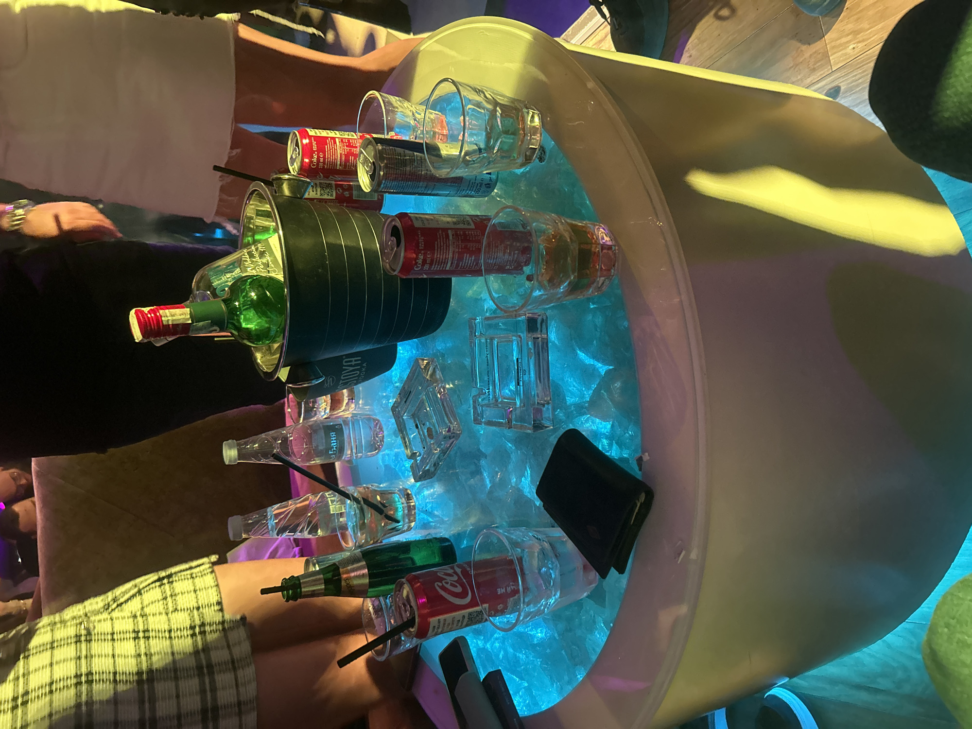 A high-angle shot depicts an ice bucket filled with ice and various drinks, including canned Coca-Cola, vodka, and bottles of water, with clear glasses nearby, some containing straws. A green bottle sits in a black holder. A black wallet is visible among the ice. Parts of people surround the bucket; a wrist with a watch, a white shirt, a plaid shirt, and other indistinct clothing can be seen. The scene is illuminated with a blue glow from within the bucket, indicating it may be a light-up ice bucket. The location and city of Veliko Tarnovo, Bulgaria cannot be determined from this image alone.