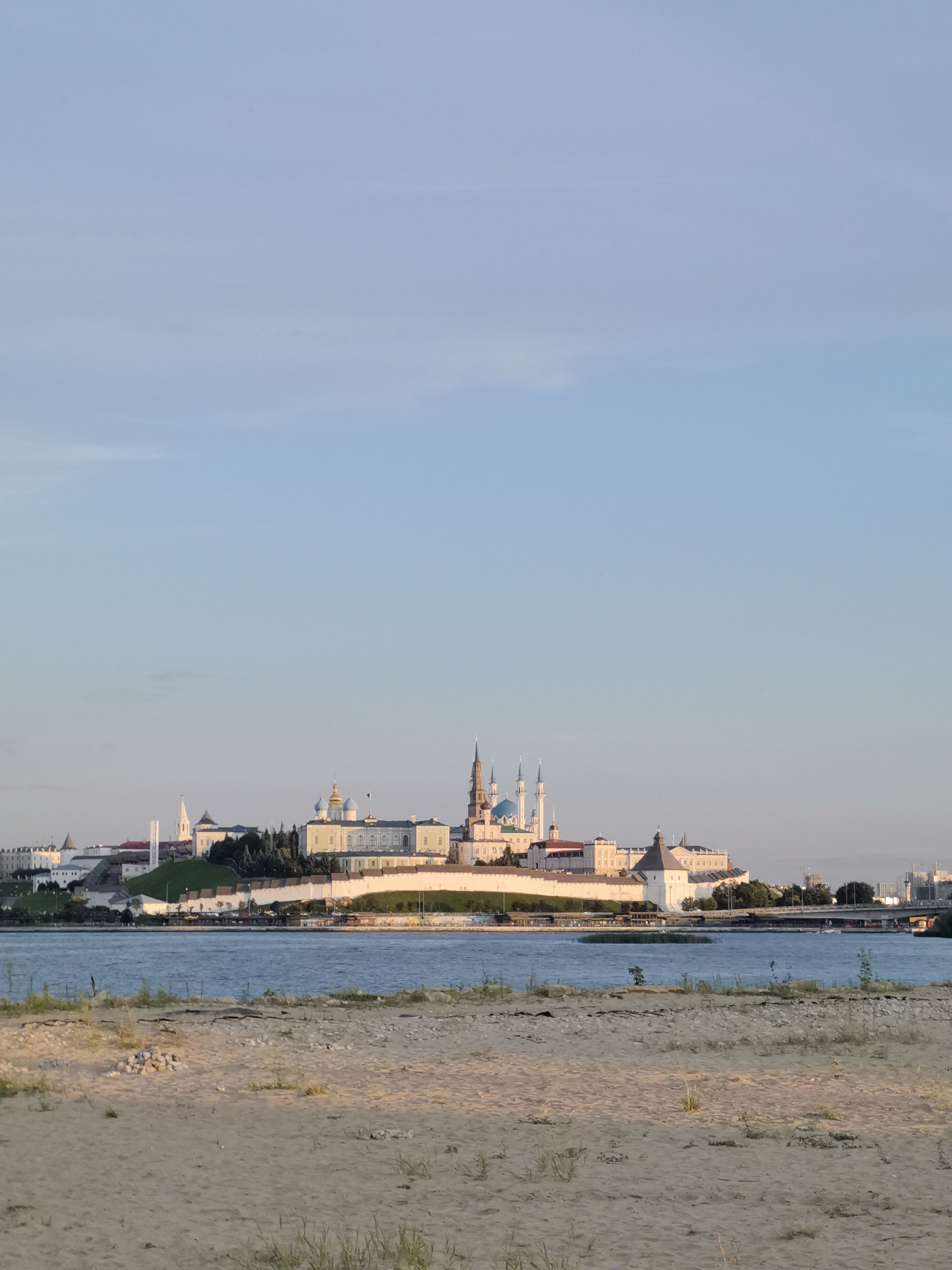 The image depicts a panoramic view of the Kazan Kremlin, a UNESCO World Heritage site, situated on a hill overlooking a body of water, likely the Kazanka River. The Kremlin's distinctive architecture, including white fortress walls, numerous towers, and several domed buildings with a prominent blue dome, is clearly visible. The scene is bathed in the warm light of either sunrise or sunset, casting long shadows and creating a serene atmosphere. In the foreground, a sandy beach with sparse vegetation leads to the water, suggesting a public access point or recreational area. Beyond the river, a modern bridge and urban skyline of Kazan can be seen in the distance, indicating the city's coexistence of historical and contemporary elements. The sky is clear and light blue, typical of a fair weather day. No people are visible in the image.