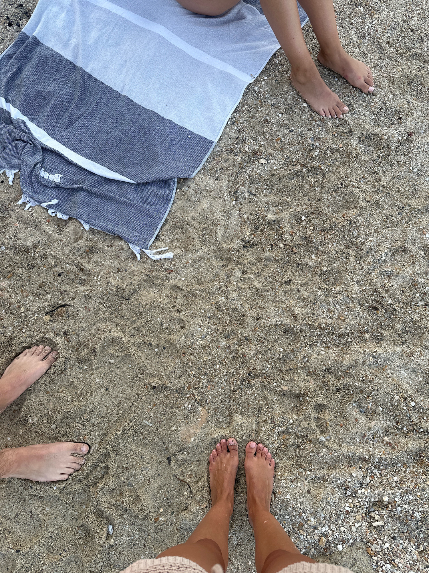 The image depicts a top-down view of a sandy beach in the Kassandra Municipal Unit, Greece. A striped beach towel, predominantly dark blue and white, is draped on the sand in the upper left portion of the frame. In the lower half of the image, the bare feet of two individuals are visible. One set of feet, on the left, appears to be a person with lighter skin and hair on their legs. The other set of feet, positioned in the lower right, belongs to a person with darker skin and painted toenails. The sand is coarse and composed of small pebbles and shell fragments. The overall scene suggests a relaxed day at the beach.