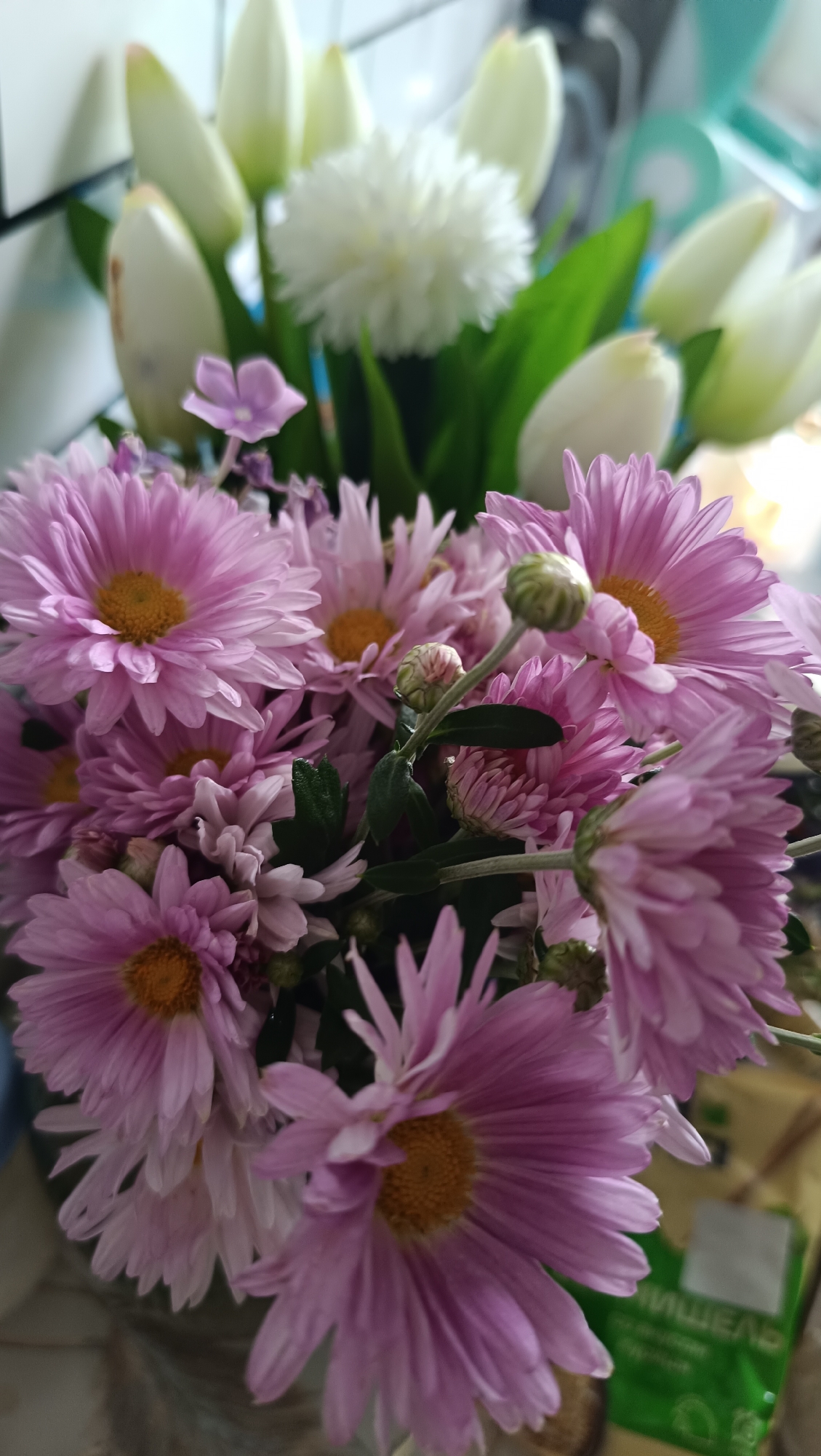 A close-up shot captures a bouquet of pink chrysanthemums with bright yellow centers, some in full bloom and others as buds. Interspersed are white tulips with green stems and leaves, along with a small, delicate purple flower. The background is softly blurred, showing hints of a light-colored wall and a green package with Russian text. The scene is likely indoors, possibly a home or shop in the city of Chaikovsky, Russia.