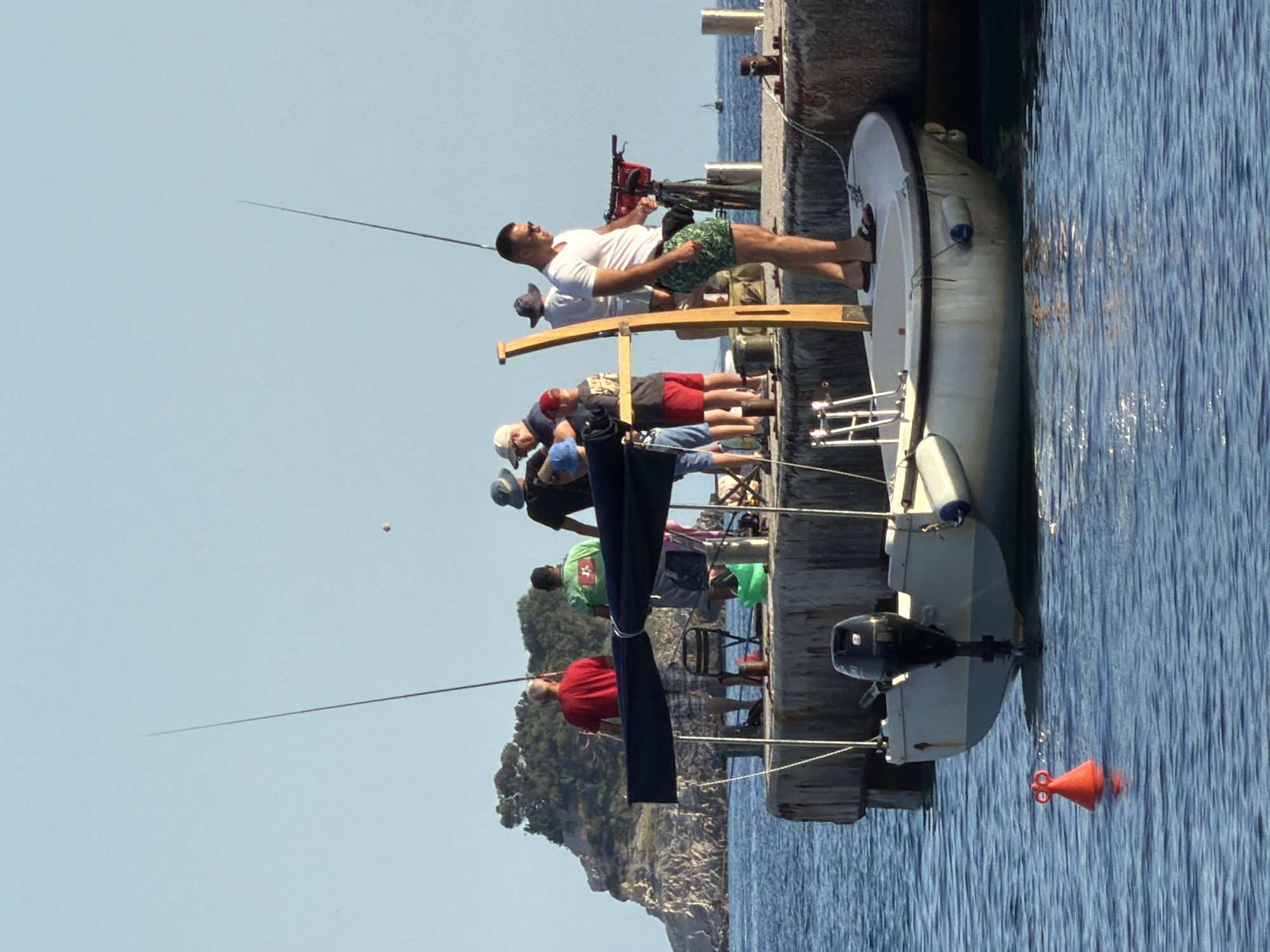 The image depicts a group of people fishing from a pier in Budva, Montenegro. The location appears to be near a rocky coastline with clear blue water surrounding the pier. Several individuals are visible, some holding fishing rods and others standing near a small white boat moored to the pier. One man stands prominently near the boat, wearing a white t-shirt and patterned shorts, and appears to be operating a red fishing machine or tool. Others are observing the scene, some seated. The sky is clear and blue, indicating a sunny day. The overall impression is one of leisure activity at a coastal location in Budva.