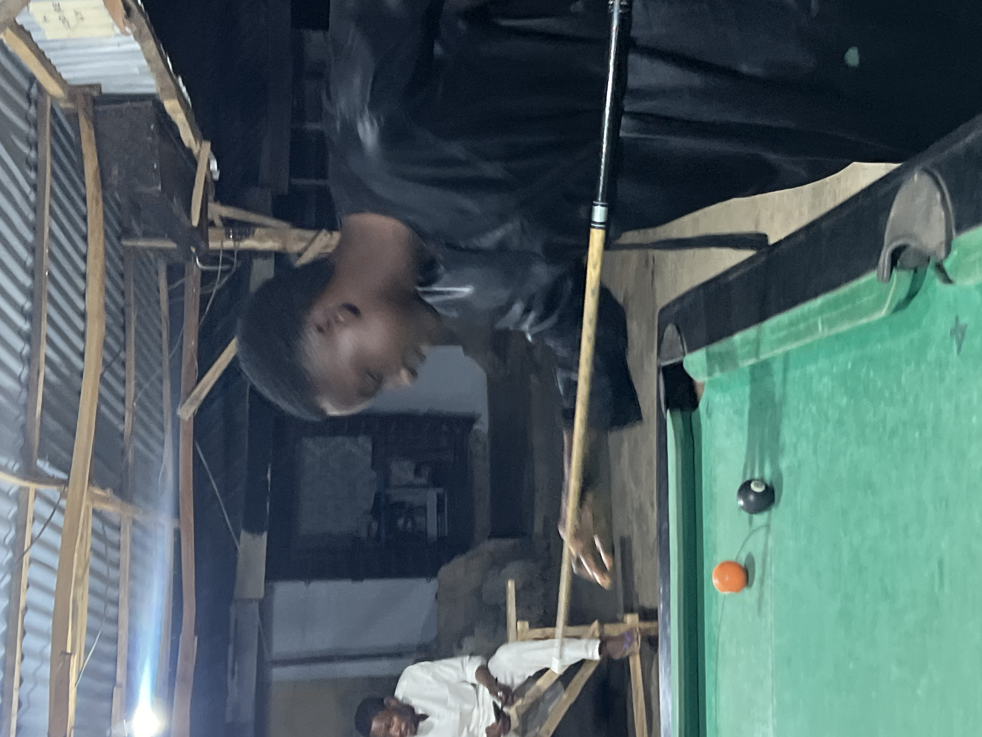 The image shows two men engaged in a game of pool within a casual, somewhat rustic setting, likely an indoor or semi-enclosed space. The photo is rotated 90 degrees clockwise.
In the foreground, a man wearing a dark, long-sleeved top is leaning over a pool table, holding a pool cue. His face is visible in profile, looking down intently at the table, suggesting he is preparing to take a shot. His left hand is poised on the cue stick. The pool table has a vibrant green felt surface, and an orange pool ball and a black 8-ball are visible on it.
In the background, another man, dressed in a white long-sleeved outfit, is seated on a wooden bench or frame, observing the game. The surrounding environment consists of visible wooden support beams and corrugated metal sheets, indicating a simple or makeshift construction style. Bright artificial lights are visible in the background, suggesting the activity is taking place at night or in a dimly lit interior. The overall scene depicts a relaxed social gathering focused on the game of pool. No discernible text is visible. This informal setting is consistent with many local community spaces found in regions like Minna, Nigeria.