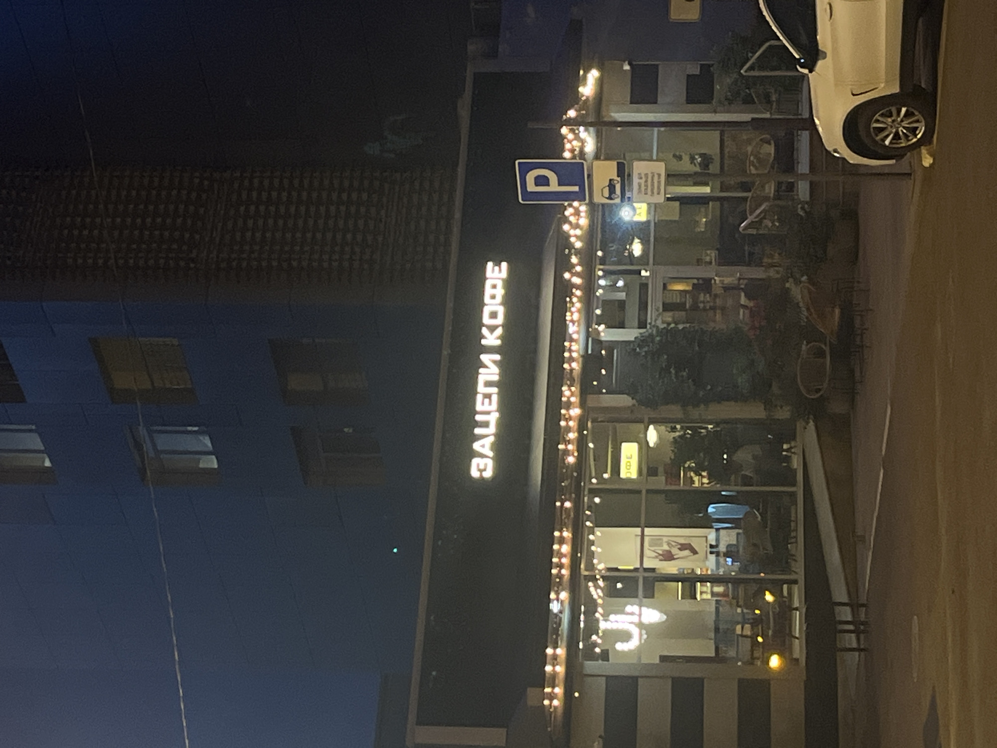 The image shows a storefront at night, likely in Krasnodar, Russia, as indicated by the Cyrillic text "ЗАЦЕПИ КОФЕ" which translates to "Hook Coffee" or "Catch Coffee". The storefront is illuminated by string lights and interior lighting, revealing a cafe with large glass windows. Inside, patrons can be seen sitting at tables. To the right of the storefront, a white SUV is parked, and a blue parking sign is visible. The building behind the cafe appears to be a multi-story structure with illuminated windows. The overall scene suggests a commercial area in a city at night.