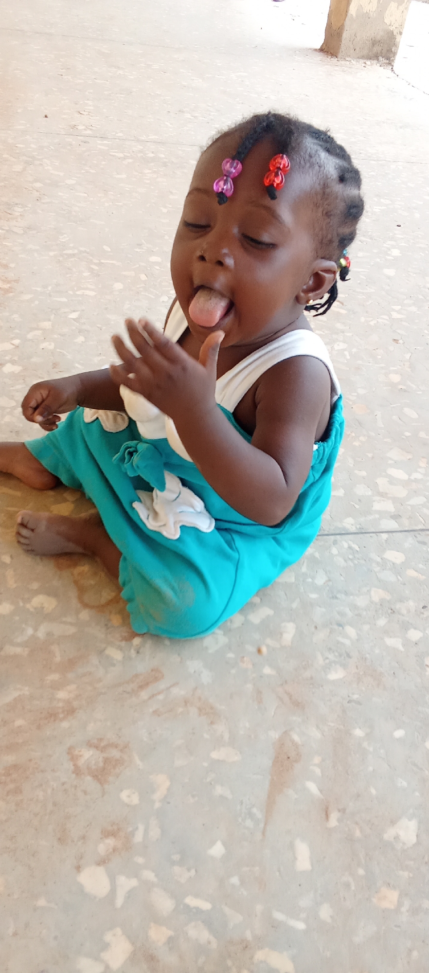 A child sits on a mottled beige floor. The child has dark skin, dark hair with colorful beads, and is wearing a blue dress with white accents. The child's eyes are closed and the tongue is protruding. One hand is raised near the face. In the background, a light beige wall is visible. The location is Akwanga, Nigeria.