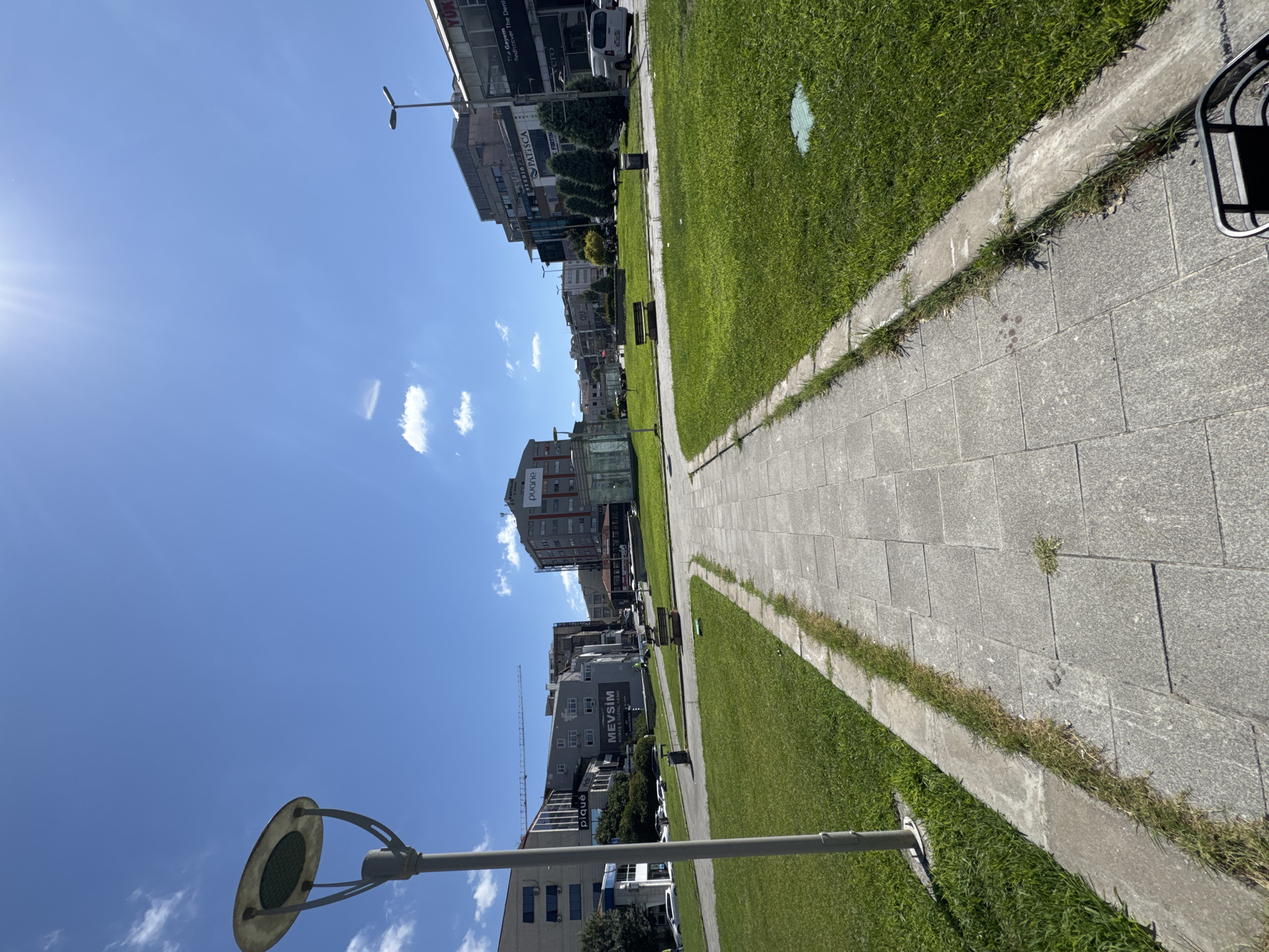 The image shows a paved walkway with a concrete border leading into a grassy park area. A single lamppost stands to the left of the walkway. In the background, there are several buildings, including a tall building with a crane next to it, and other mid-rise buildings. Some of the buildings have signs visible, such as "pique," "MEVSIM," "PUANE," and "YUKSEK." The sky is bright blue with a few white clouds and the sun is shining brightly. This scene is located in Gungoren, Turkey.