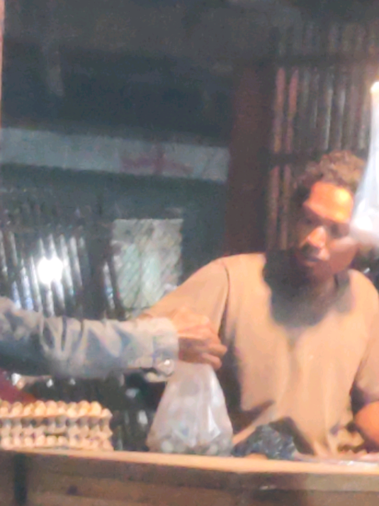 The image shows a close-up of a transaction at a market stall. A person's arm and hand are visible on the left, holding a clear plastic bag filled with what appear to be coins. They are handing the bag to another person, whose face is partially visible in the center of the frame. This person, likely a vendor, has dark skin and is wearing a light-colored t-shirt. Their face is illuminated by a bright light source from above and to the right. Behind the vendor, a corrugated metal wall is visible. In the foreground, a tray of eggs is partially visible on a wooden counter. The overall setting suggests a marketplace in Karawang, Indonesia. The lighting is dim and the image quality is low, with significant noise and blur, particularly in the background elements and the faces of the individuals.