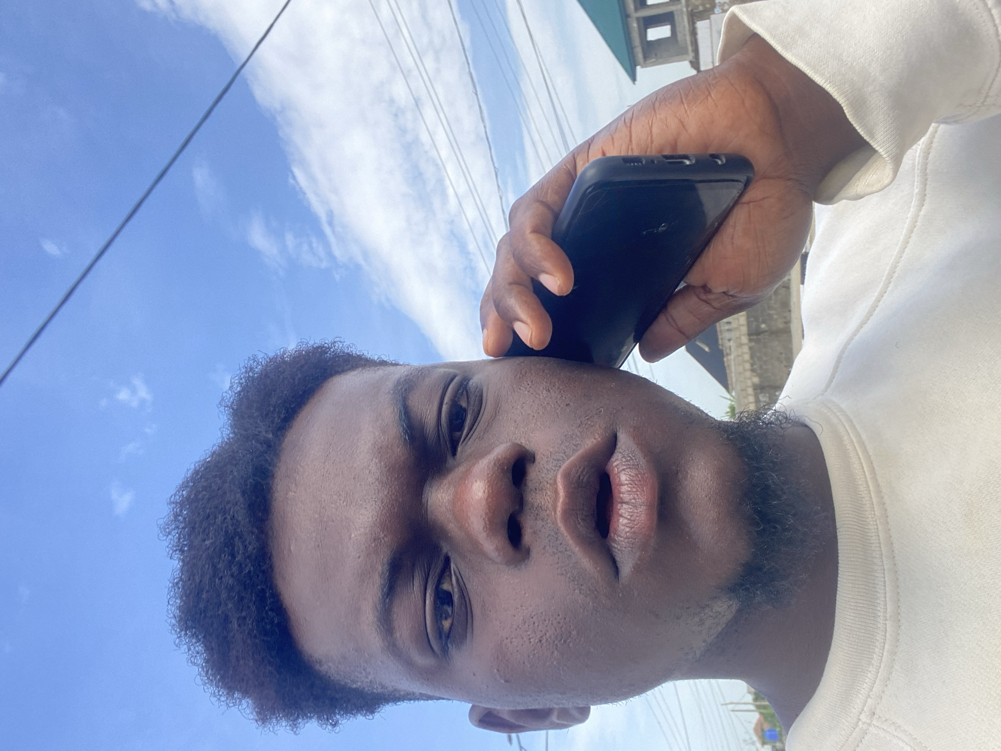 A low-angle, close-up portrait of a dark-skinned man holding a black smartphone to his ear. The man has curly black hair, a short beard, and is wearing a white sweater. The background features a partly cloudy sky and utility lines, with a building partially visible in the upper right corner. The location is Unknown, Unknown.
