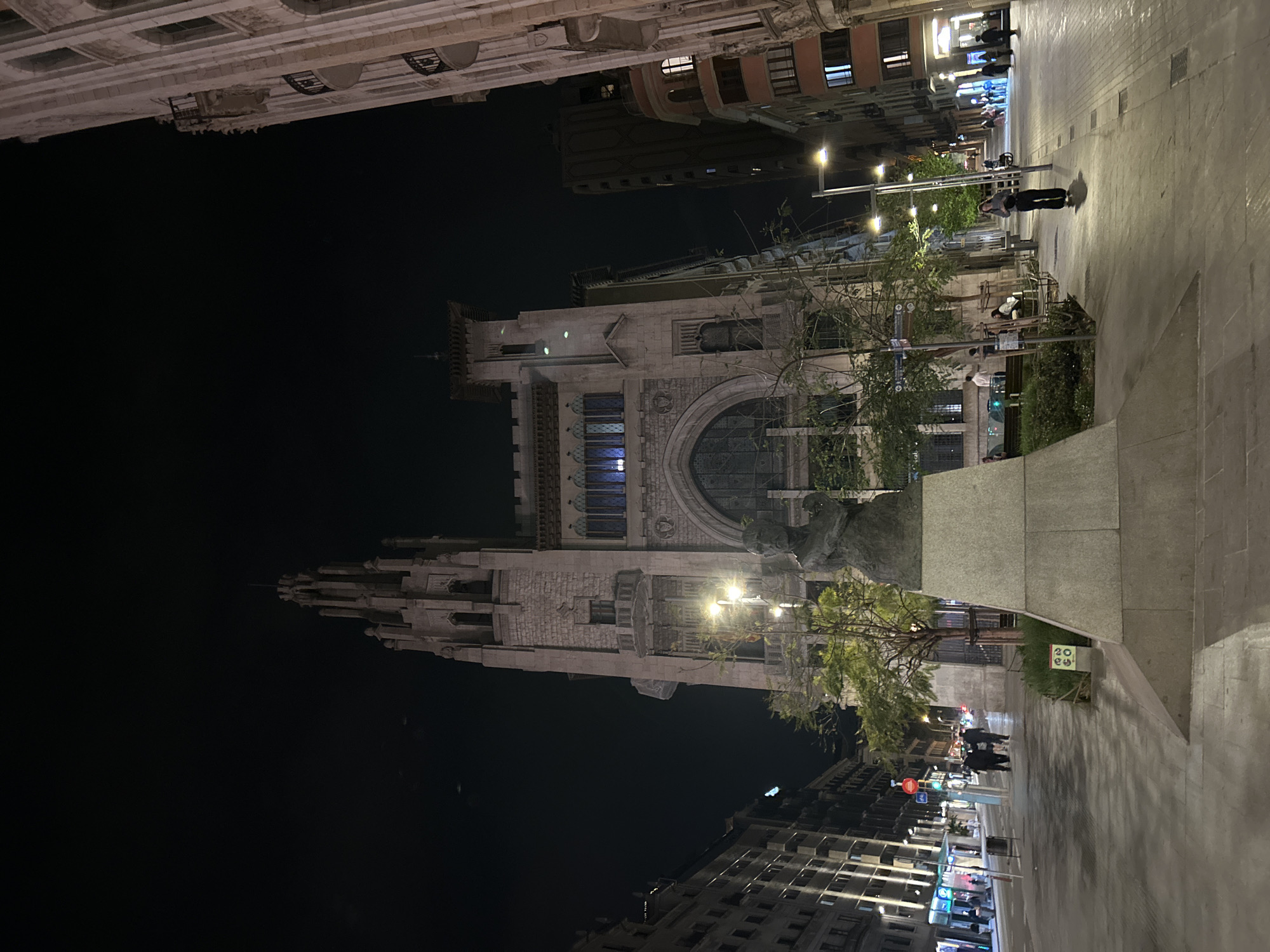 The image depicts a night scene in Barcelona, Spain, showcasing the Ajuntament de Barcelona (Barcelona City Hall) from a ground-level perspective. The Gothic-style architecture of the building is illuminated, contrasting against the dark sky. Streetlights line the pedestrian walkway, casting light on the pavement and nearby buildings. People can be seen walking along the street. A statue is visible in front of the city hall. Other buildings line the street.