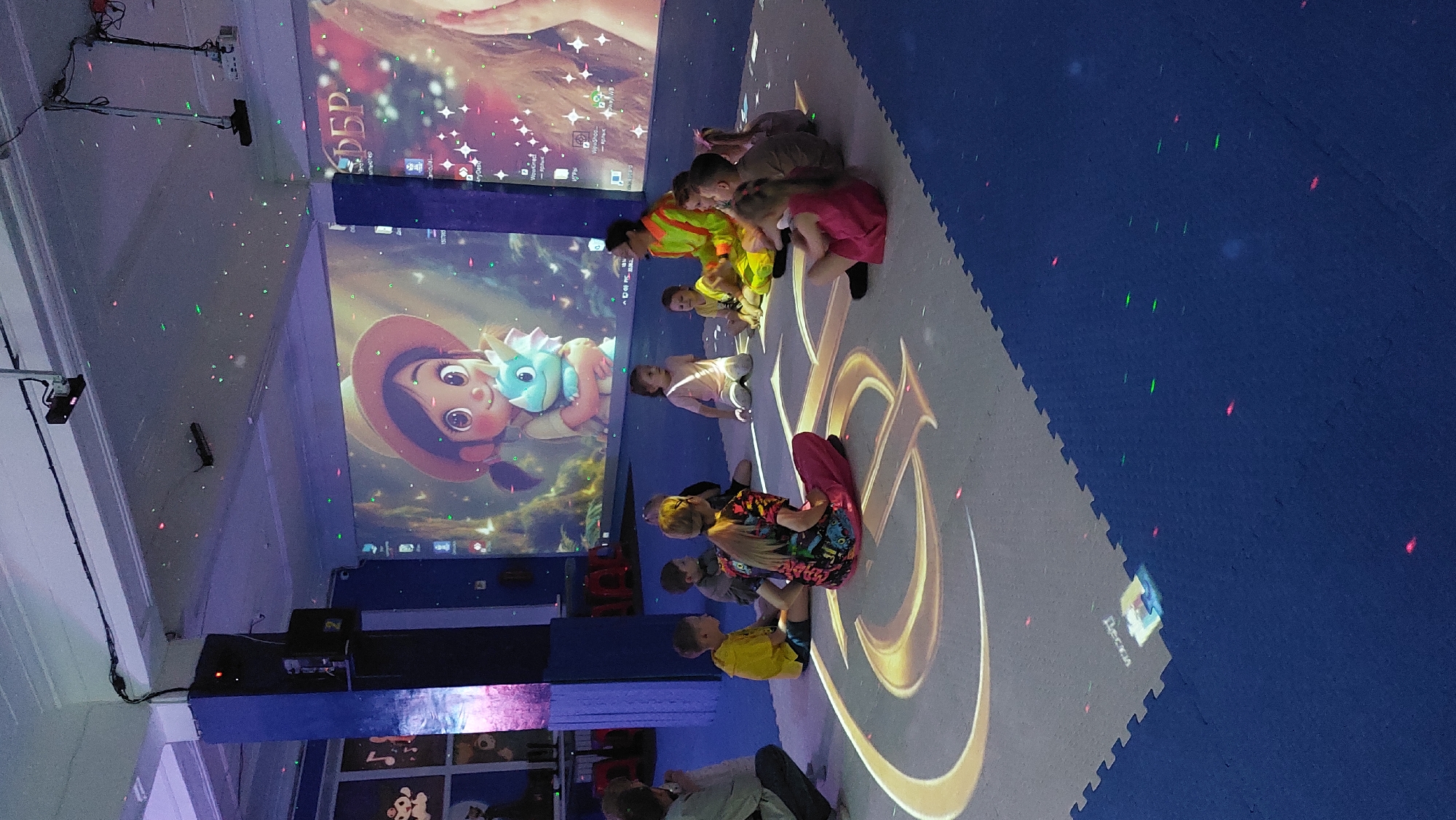 A group of children are seated on a blue padded floor in front of a large projection screen. The projection shows a colorful animated scene of a young girl with a blue dragon-like creature. Scattered green and red dots are projected onto the floor and ceiling, resembling small lights or stars. Some of the children are looking at the screen, while others are interacting with each other. One adult, wearing a bright orange and yellow vest, appears to be supervising the children. The room is dimly lit, with the projections and small lights being the primary sources of illumination. The floor is made of interlocking puzzle mats, and a large golden graphic is projected onto the floor in front of the children. A small inscription in Russian, "Дети" (Deti), meaning "Children," is visible on the floor projection. The overall scene suggests a playful and engaging environment, possibly a children's play area or educational center. The time of day is not evident, and there are no clear weather cues. The emotions of the children appear to be calm and attentive.