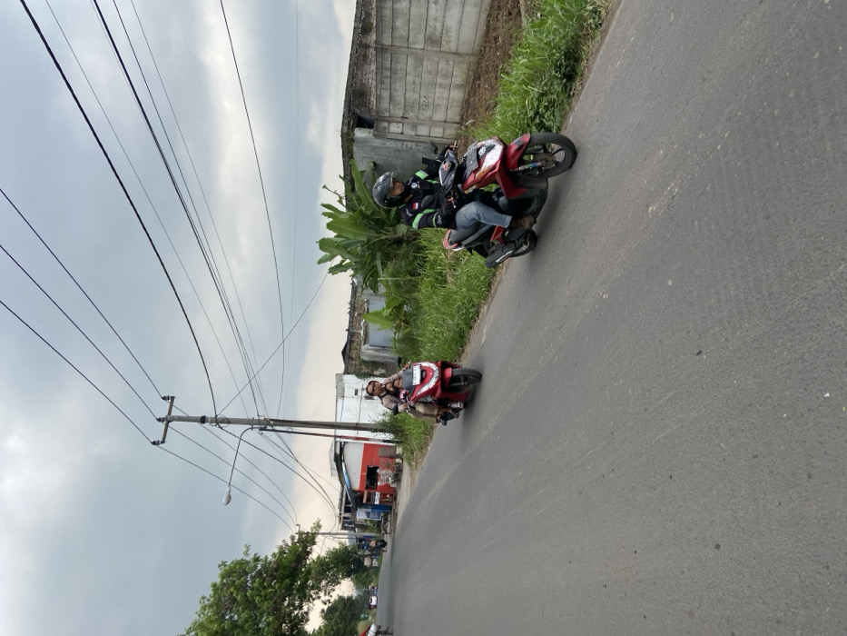 Two men are riding red motorcycles on a paved road in Kecamatan Margaasih, Indonesia. The road is lined with grass and trees. The sky is overcast with gray clouds. The men are both wearing helmets. One man is wearing a red and white jacket. The other man is wearing a light brown shirt. The motorcycles are both facing the same direction and are travelling on the right side of the road. The road appears to be empty of other traffic. There are multiple power lines overhead. The location is approximately -6.963 latitude and 107.538 longitude.