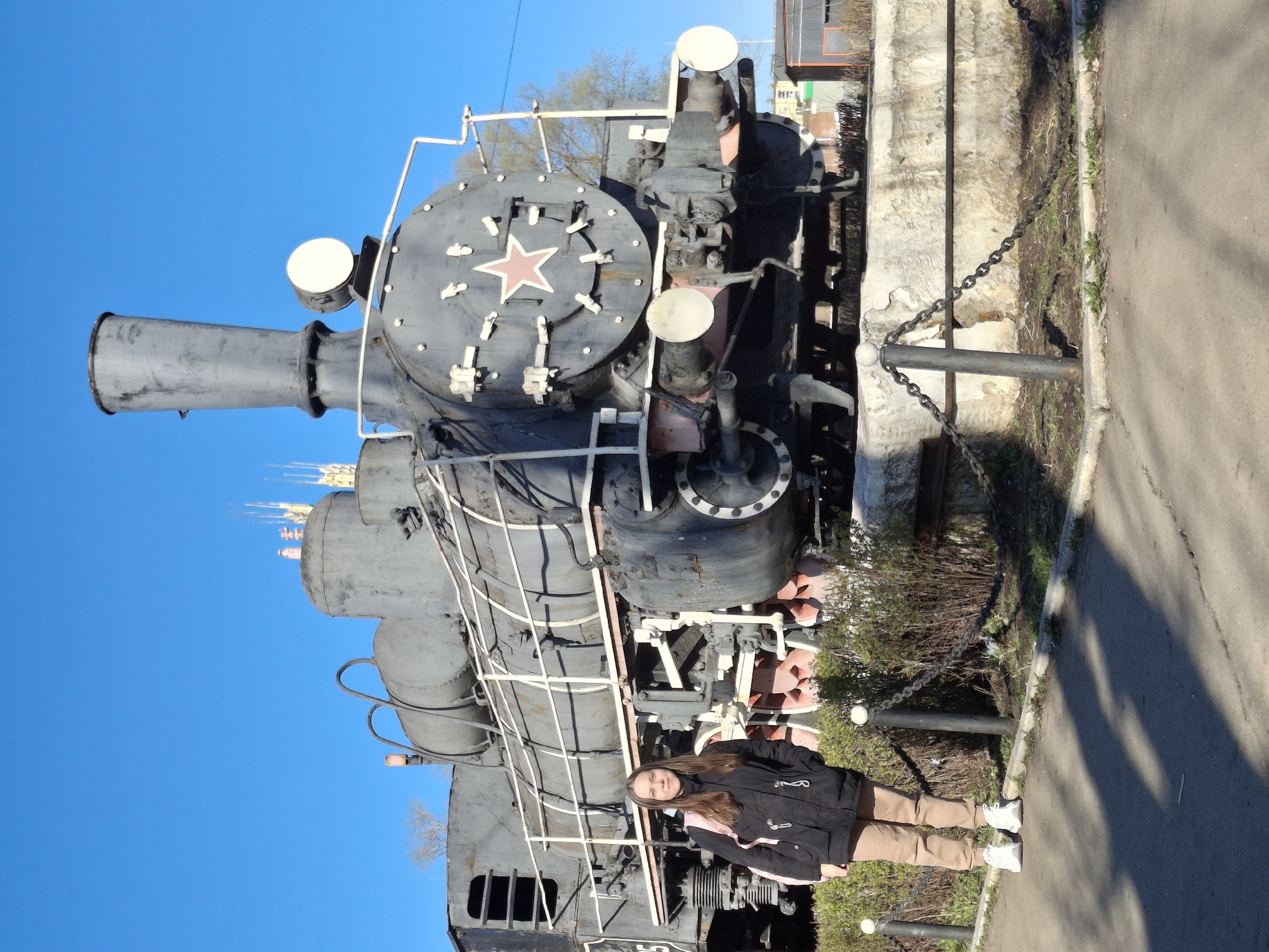 The image shows a woman standing next to a large, old steam locomotive. The locomotive is gray, with a prominent red star on its front. The locomotive is located on a concrete platform bordered by a chain fence. The sky is bright blue. The woman is wearing a dark jacket and light brown pants, and white sneakers. The photo was taken in Sarapul, Russia.