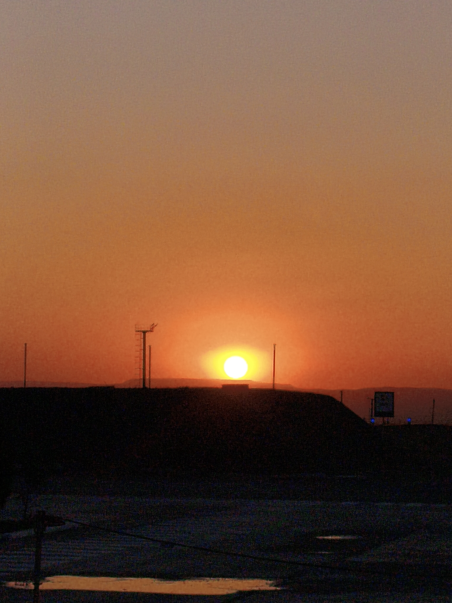The image depicts a sunset over a dark, silhouetted landscape. The sky is a gradient of orange and pink, with the sun appearing as a bright, yellow orb positioned slightly to the right of the center of the frame. In the foreground, a dark, hilly silhouette suggests a natural terrain or an elevated structure. On the left side of the silhouette, a tall, thin pole is visible. To the right of the sun, a prominent structure resembling a communications tower with visible rungs or ladders stands against the bright sky. Further to the right, a square sign with a darker blue inset and some text is mounted on a pole, with two small blue lights visible below it. The foreground below the silhouette is a dark, textured area, possibly indicating a ground surface or water. The overall impression is one of a remote, possibly industrial or airport-like setting at dusk.