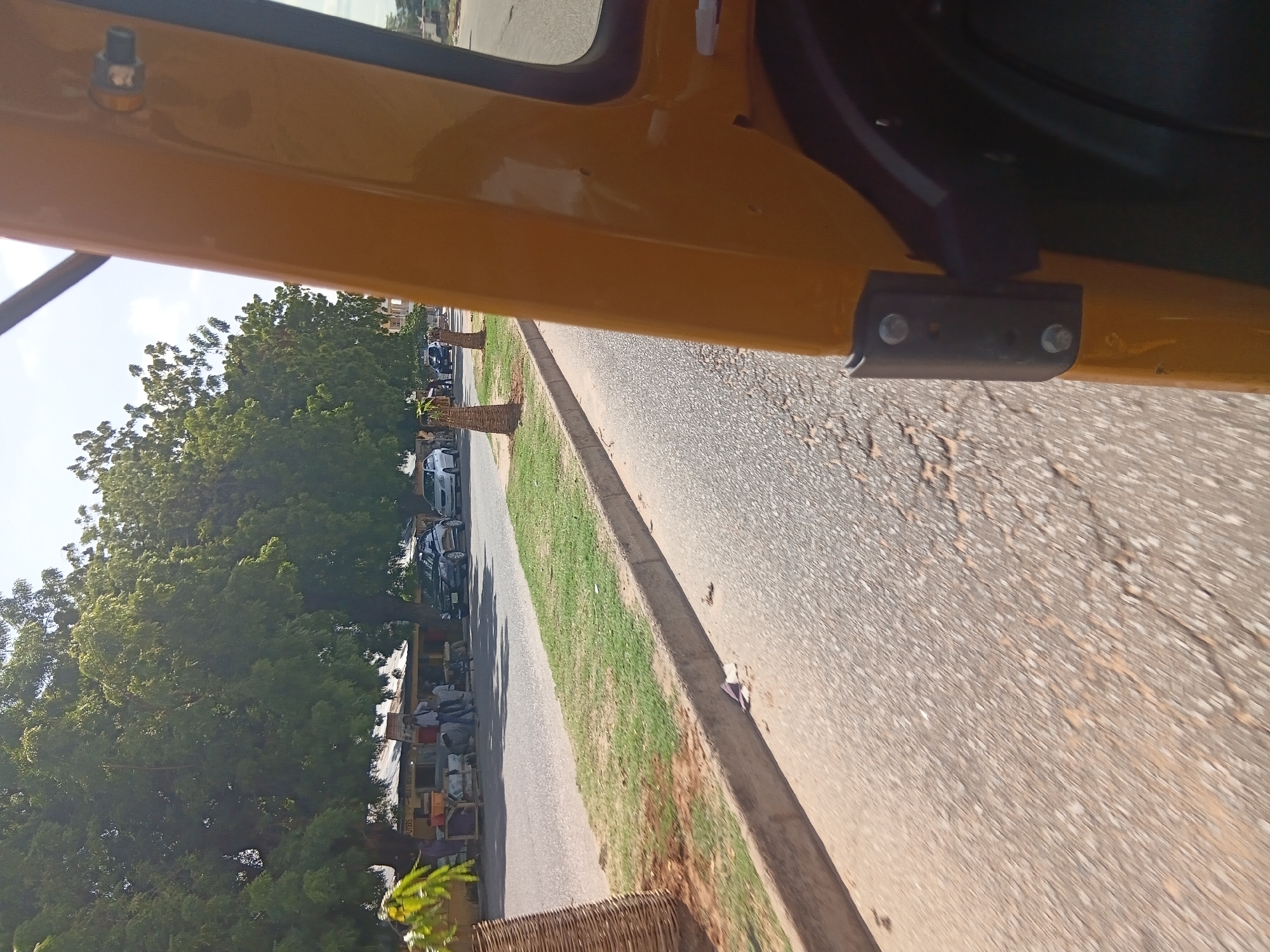 The image shows a view from inside a yellow vehicle, likely a tuk-tuk, traveling along a road in Jalingo, Nigeria. On the left side of the road, there is a row of shops with corrugated metal roofs, shaded by large, leafy green trees. Several people are visible standing and walking in front of the shops. A few cars are parked or driving on the road. The road itself appears to be paved with asphalt and shows signs of wear. A grassy median strip separates the road from the sidewalk in front of the shops. The sky is bright and clear.