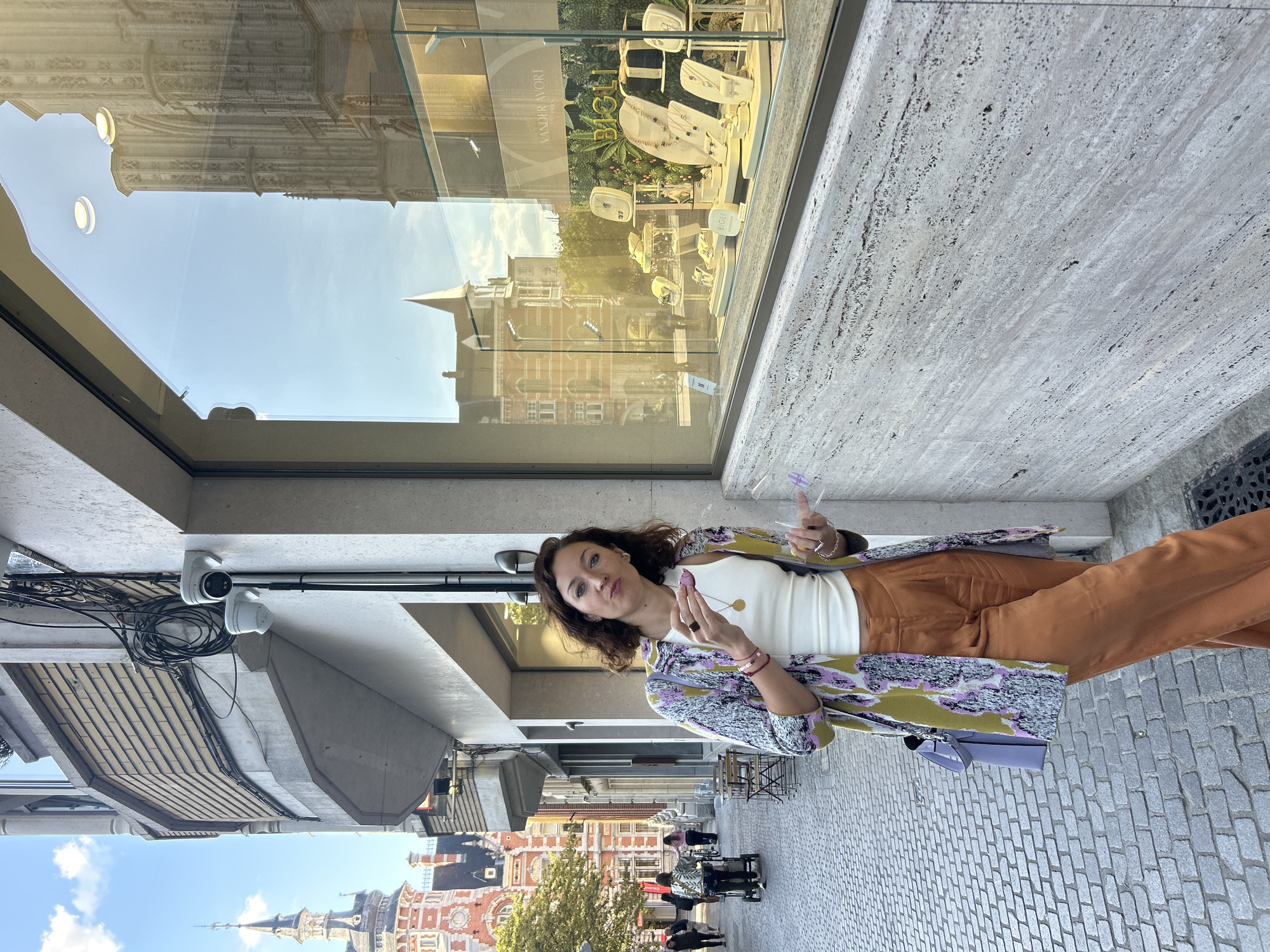 A woman stands on a cobblestone street in Leuven, Belgium, in front of a jewelry store window. The woman is wearing a white tank top, orange wide-leg pants, and a colorful patterned jacket. She is holding a small purple object in her right hand and a clear plastic object in her left hand, and appears to be looking at the camera with a slight smile. The jewelry store window displays various necklaces, earrings, and rings. In the background, buildings with distinctive architecture, a church spire, and trees can be seen. The sky is blue with some clouds. The scene suggests an outdoor urban setting during the daytime.