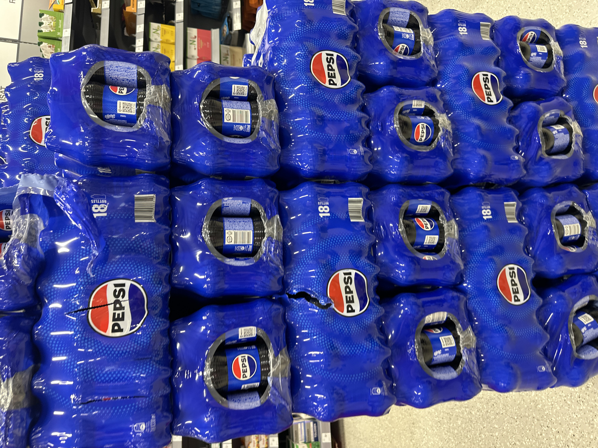 This media file is a close-up shot of stacked packs of Pepsi bottles. The packs are predominantly blue with the iconic red, white, and blue Pepsi logo clearly visible on several of them. The bottles themselves are also visible through cutouts in the packaging. The packs appear to be shrink-wrapped, and some have labels indicating "18 BOTTLES". The lighting suggests an indoor setting, likely a store or warehouse, with the bright fluorescent lights casting reflections on the plastic packaging. There are no people visible in the image. The arrangement of the packs suggests they are part of a larger display or inventory. Some text like "TOLONEN" is also partially visible in the background, suggesting it might be a retail environment. The overall impression is one of commercial product display.