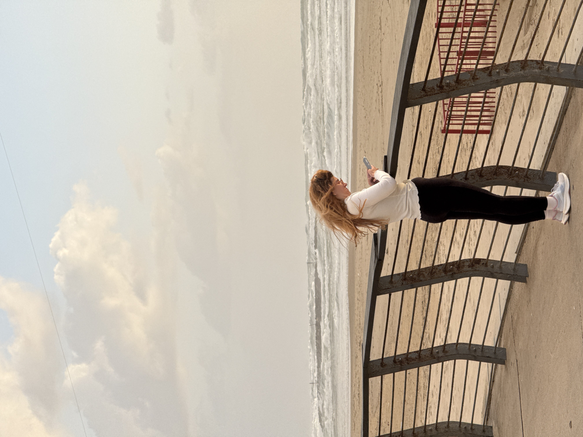 A woman stands on a promenade overlooking the beach in Tel Aviv, Israel. She is dressed in black leggings and a white long-sleeved shirt, and she is holding a cell phone, possibly taking a photo. She is standing next to a metal railing. The beach and waves are visible in the background. The sky is overcast.