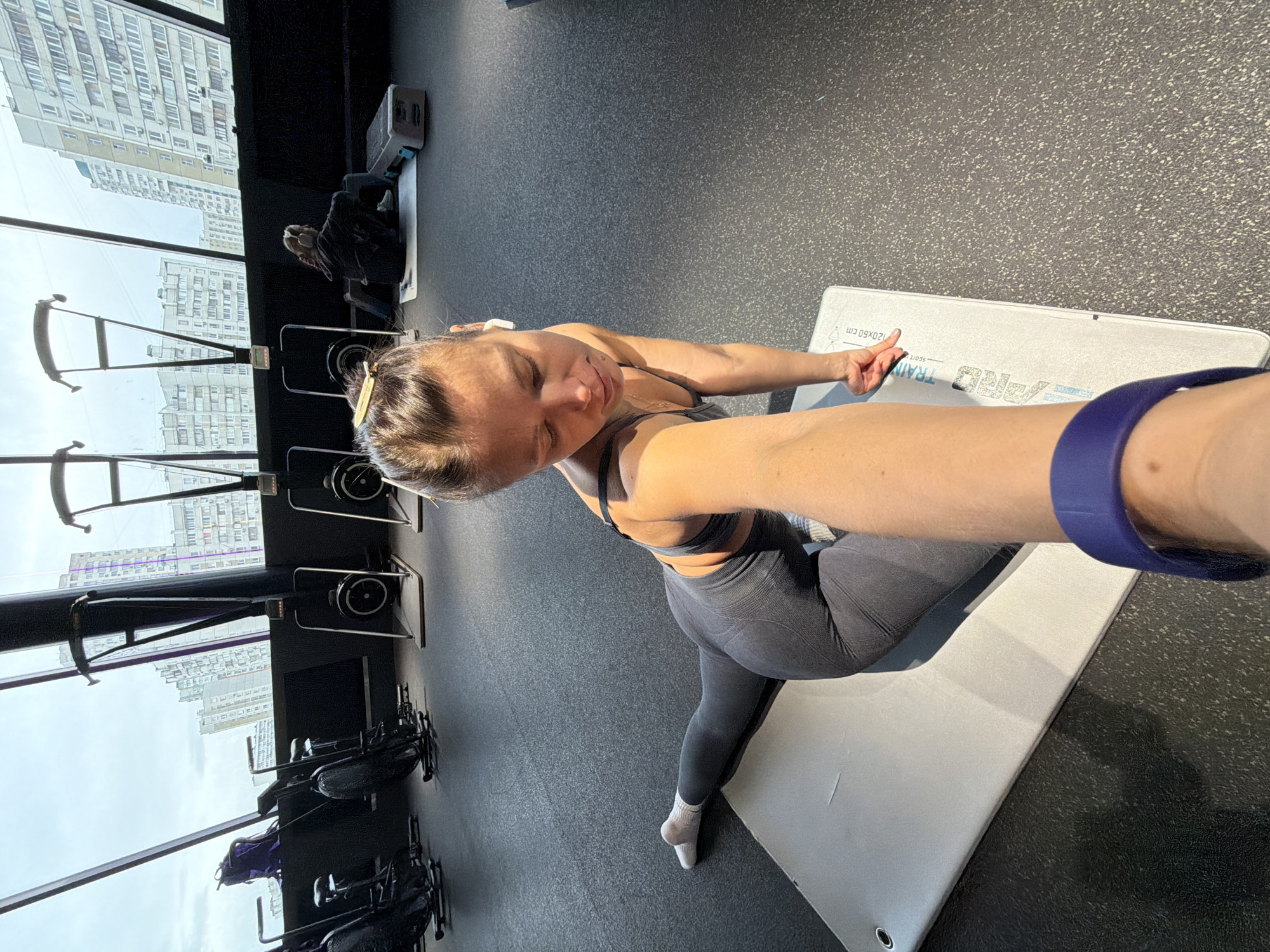 A woman in workout attire takes a selfie in what appears to be a gym. She is kneeling on a gray mat with her left arm extended, possibly holding a camera phone. She is wearing a gray sports bra and leggings, and a dark blue fitness tracker is visible on her wrist. A white wireless earbud is in her left ear. Exercise equipment, including rowing machines, is visible in the background. Large windows reveal a cityscape with tall residential buildings, indicating an urban location. Given the architectural style of the buildings, the scene may be in Moscow, Russia. The floor is a dark speckled rubber material commonly found in gyms. The light is bright, likely from the large windows. Another person is visible in the background seated on one of the rowing machines.