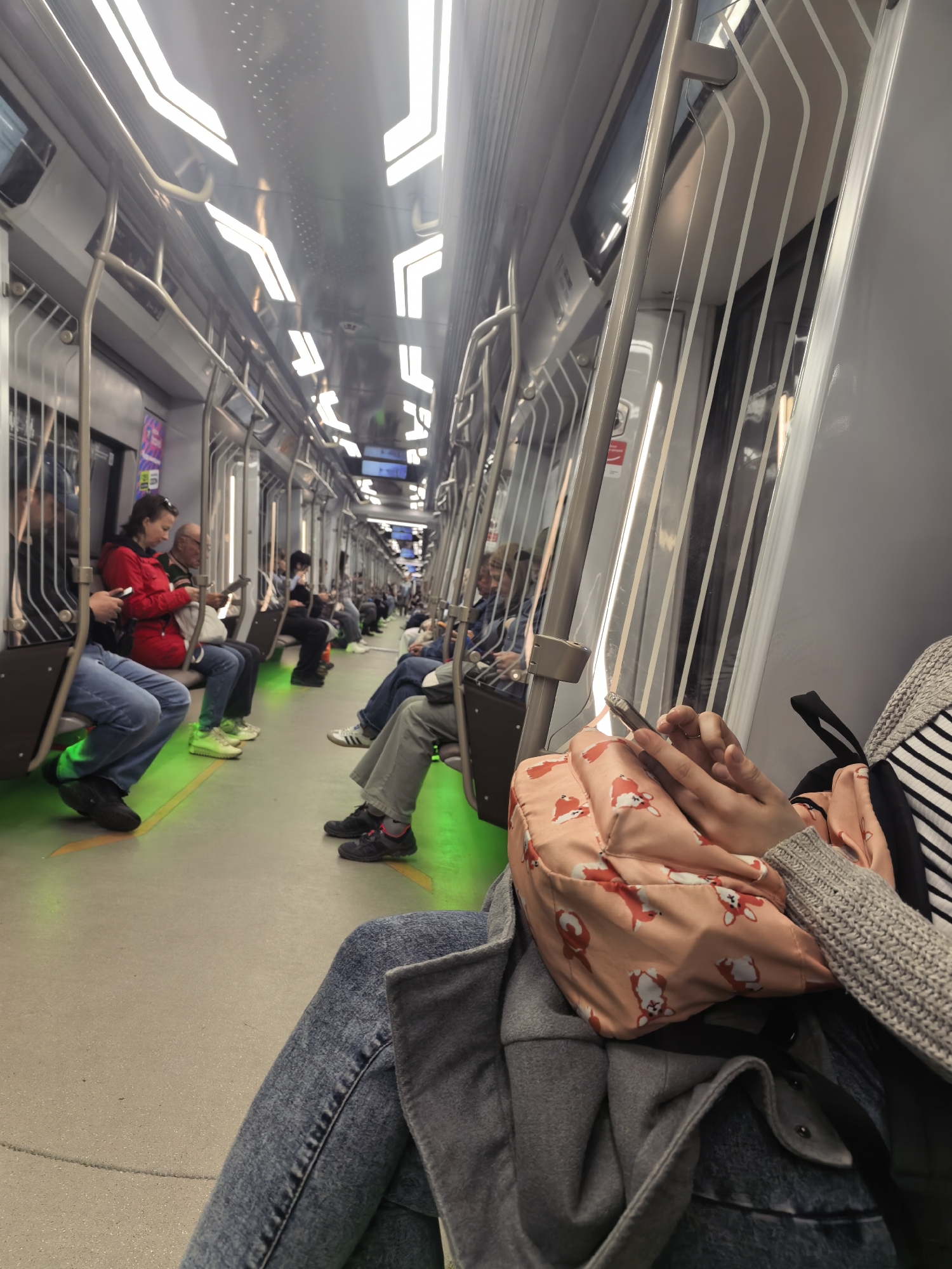 The interior of a subway car is visible, filled with passengers seated along the sides. The car's design features a metallic frame with vertical white stripes on the walls. Overhead, the ceiling is lit by a series of modern, rectangular light fixtures. Passengers are engaged in various activities, such as looking at their phones, reading, or simply sitting. The car floor glows with a green hue. A person in the foreground is looking at a smartphone. The location is a subway car in an unidentified city.