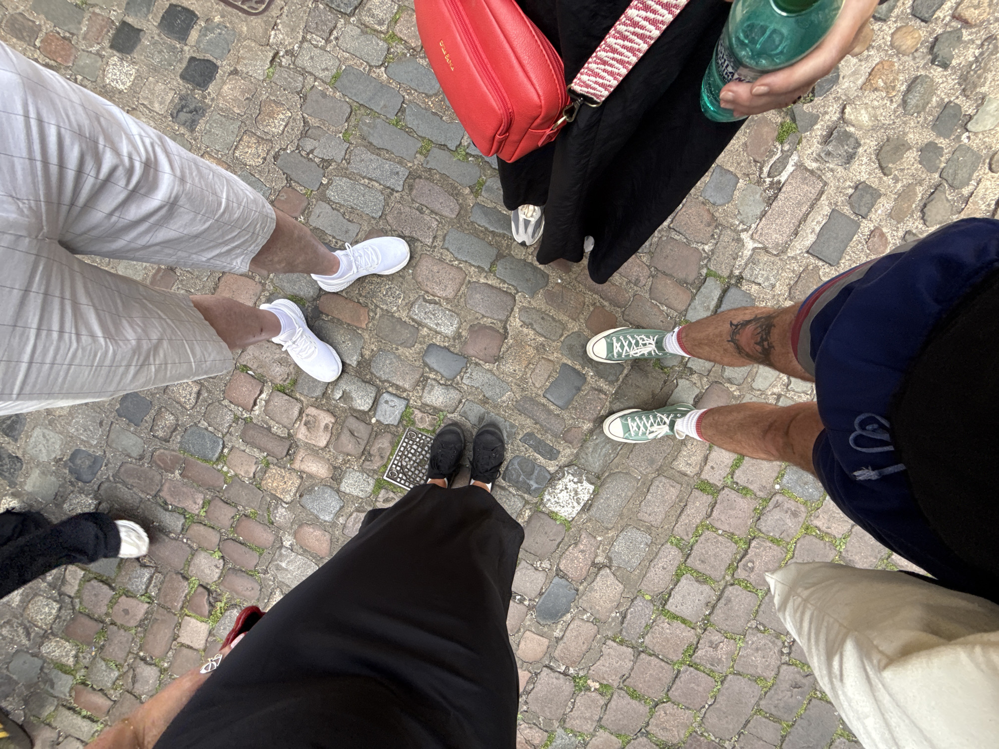 The image shows a group of five people standing in a circle on a cobblestone street. The location is in the city of Unknown, Unknown. The group is positioned close together, with their feet forming the perimeter of the circle.
Clothing and accessories:
- One person is wearing white shorts and white sneakers.
- Another person is wearing dark shoes and a long black skirt.
- Another person is wearing green sneakers, blue shorts, and a red and white striped sock.
- A red bag is also visible.
- Another person is wearing black and white sneakers and black pants.
- Another person is wearing a light-colored pants.
- Someone is holding a green glass bottle.
Environment:
- The cobblestone street is made up of stones of various sizes and colors, creating a textured surface.
- Some greenery is growing between the stones.
- A small square metal grate is visible on the cobblestone street.