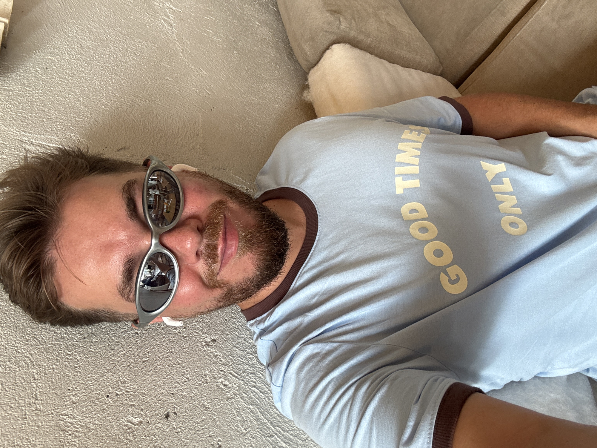 A man is captured in a selfie, lying down indoors in North Kuta, Indonesia. He has light brown hair and a neatly trimmed beard, looking directly towards the camera. He wears stylish silver-framed sunglasses that reflect an indoor setting with what appears to be furniture or shelving. A white earbud is visible in his left ear.
He is dressed in a light blue t-shirt with a brown trim on the collar and sleeves. Bold, cream-colored text on the shirt clearly reads "GOOD TIMES ONLY". The man's expression is relaxed and slightly smiling, with a content demeanor.
The setting appears to be a casual indoor space, possibly a living room or a relaxed area. Behind him, there's a neutral-toned, textured wall or floor, and plush, light-colored cushions from a couch or sofa are visible, further indicating a comfortable lounging environment. The lighting suggests it is daytime.