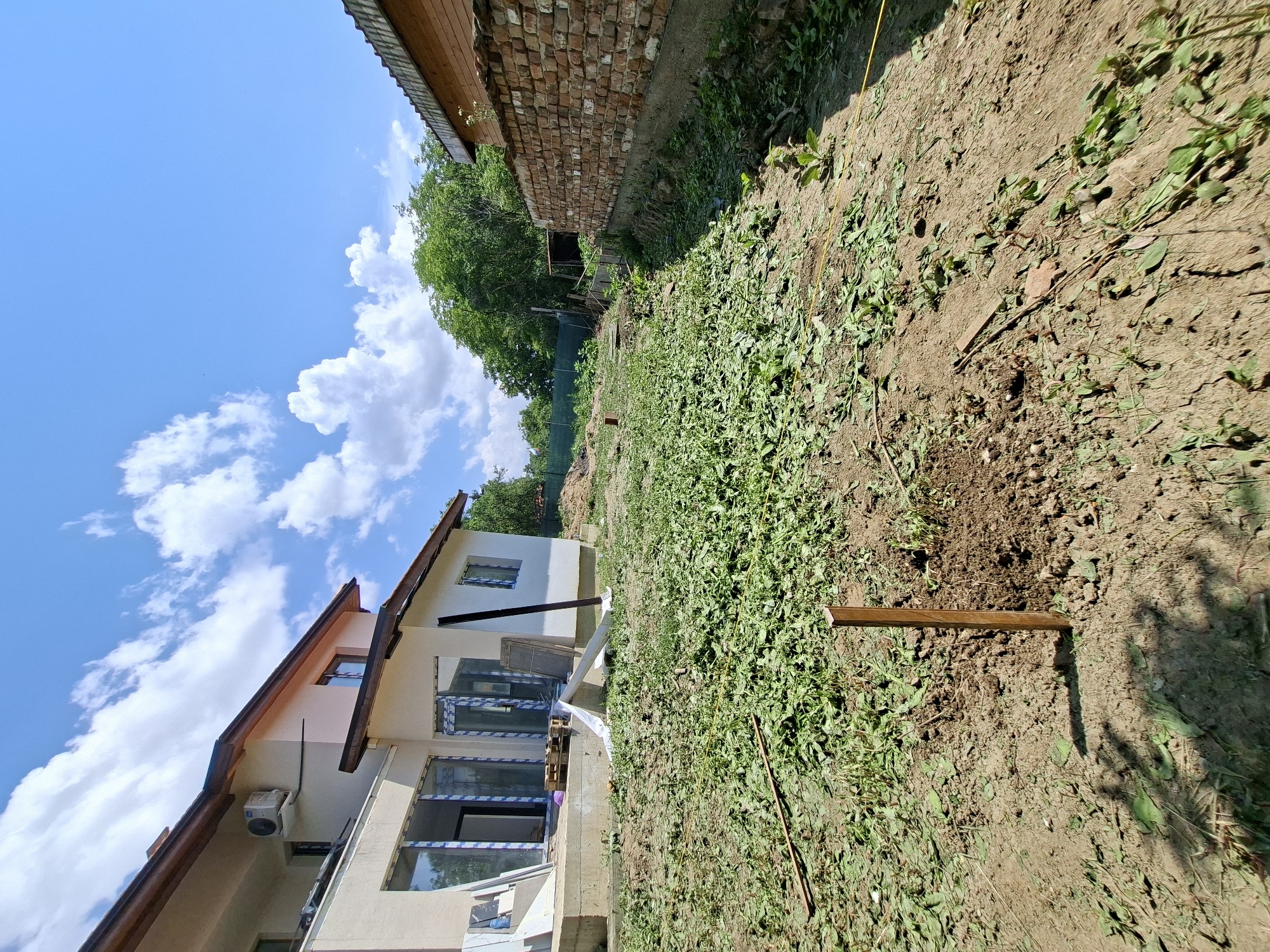 In Dolna Malina, Bulgaria, an exterior shot captures a residential construction site under a clear blue sky with scattered white clouds. In the foreground, a patch of freshly cut grass and weeds covers the uneven ground, with a wooden stake hammered into the dirt. A yellow string runs across the ground, possibly marking a boundary. To the left, a modern house with beige walls and a brown roof is visible, featuring large glass doors and windows, some of which are covered with protective plastic. An air conditioning unit is mounted on the exterior wall. Construction materials, including wooden pallets and white sheeting, are piled near the house's foundation. To the right, a brick wall with a concrete base forms a boundary, and a small wooden structure is visible behind it. Lush green trees and foliage border the property, suggesting a rural or semi-rural setting. The scene depicts an ongoing construction or landscaping project.